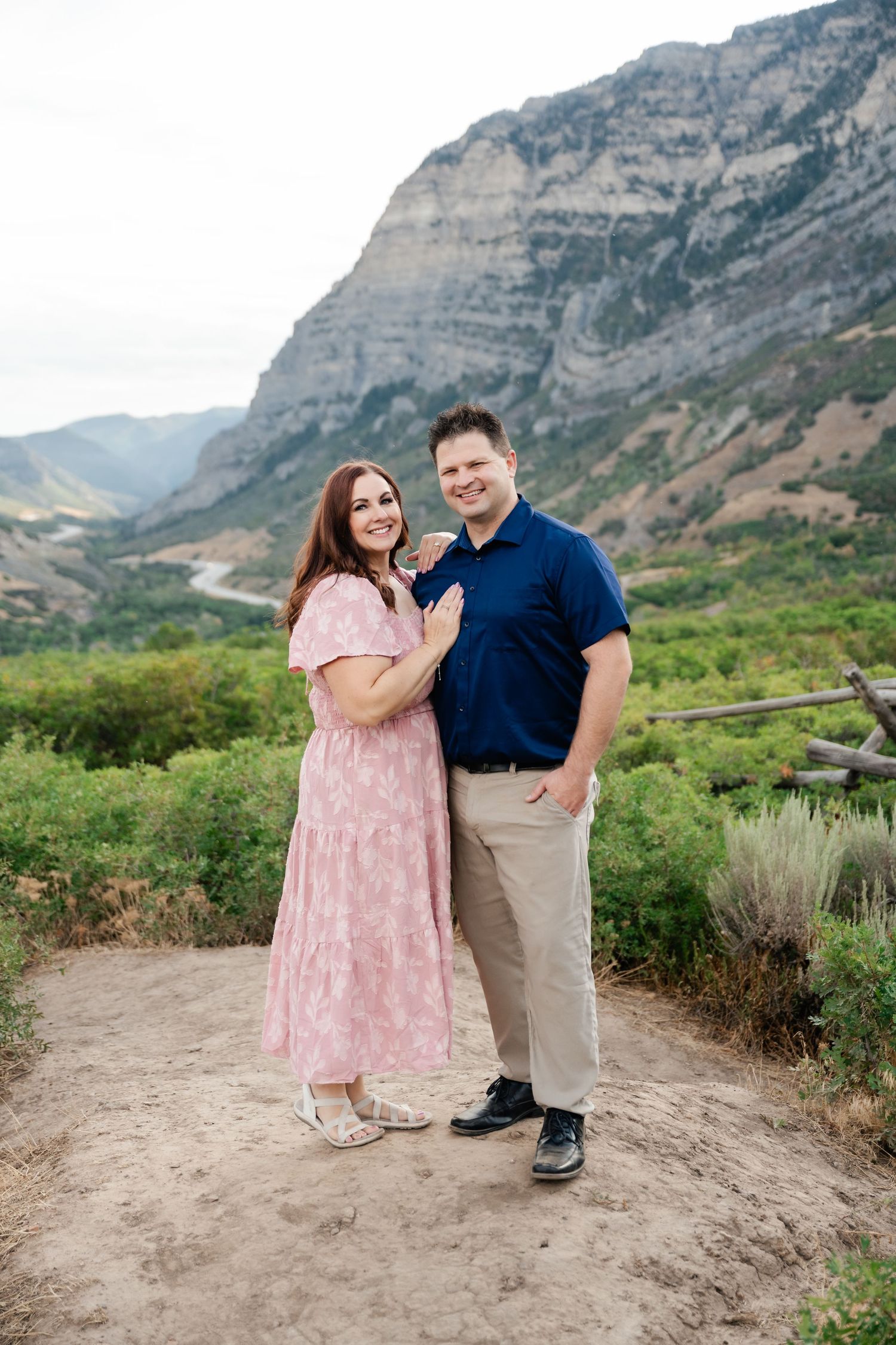 A couple poses together on a hiking trail with dramatic mountain cliffs and scenic valley views in the background.