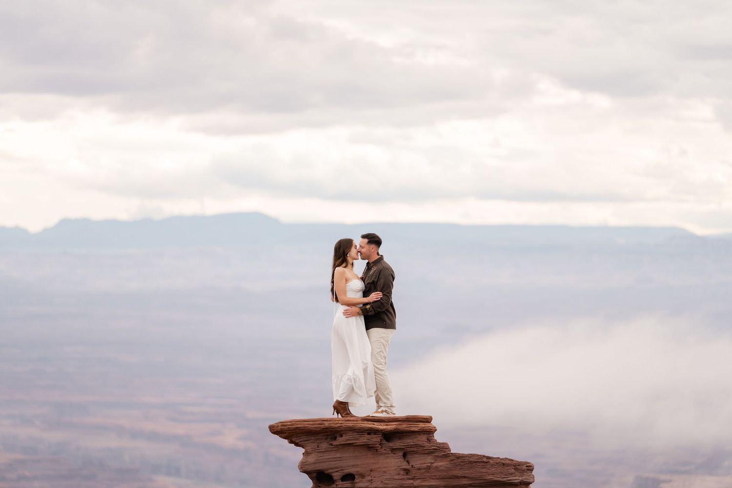 A couple shares an intimate moment on a dramatic cliff edge overlooking misty mountain vistas at sunset.