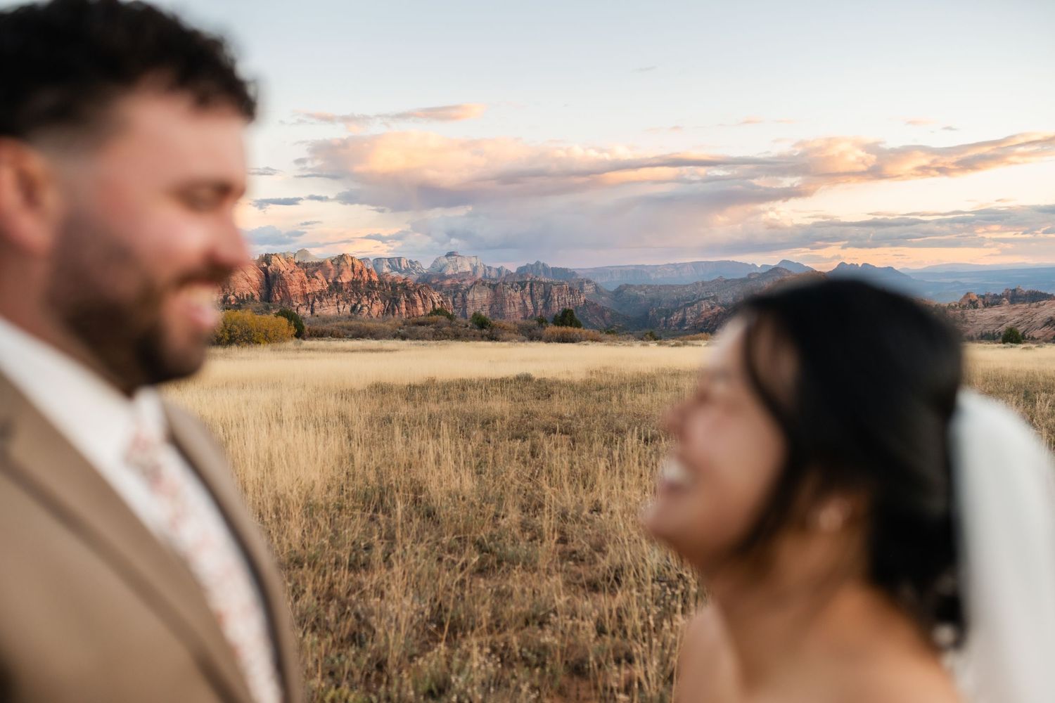 A couple embraces in a golden field at sunset with mountains and city skyline in the background.