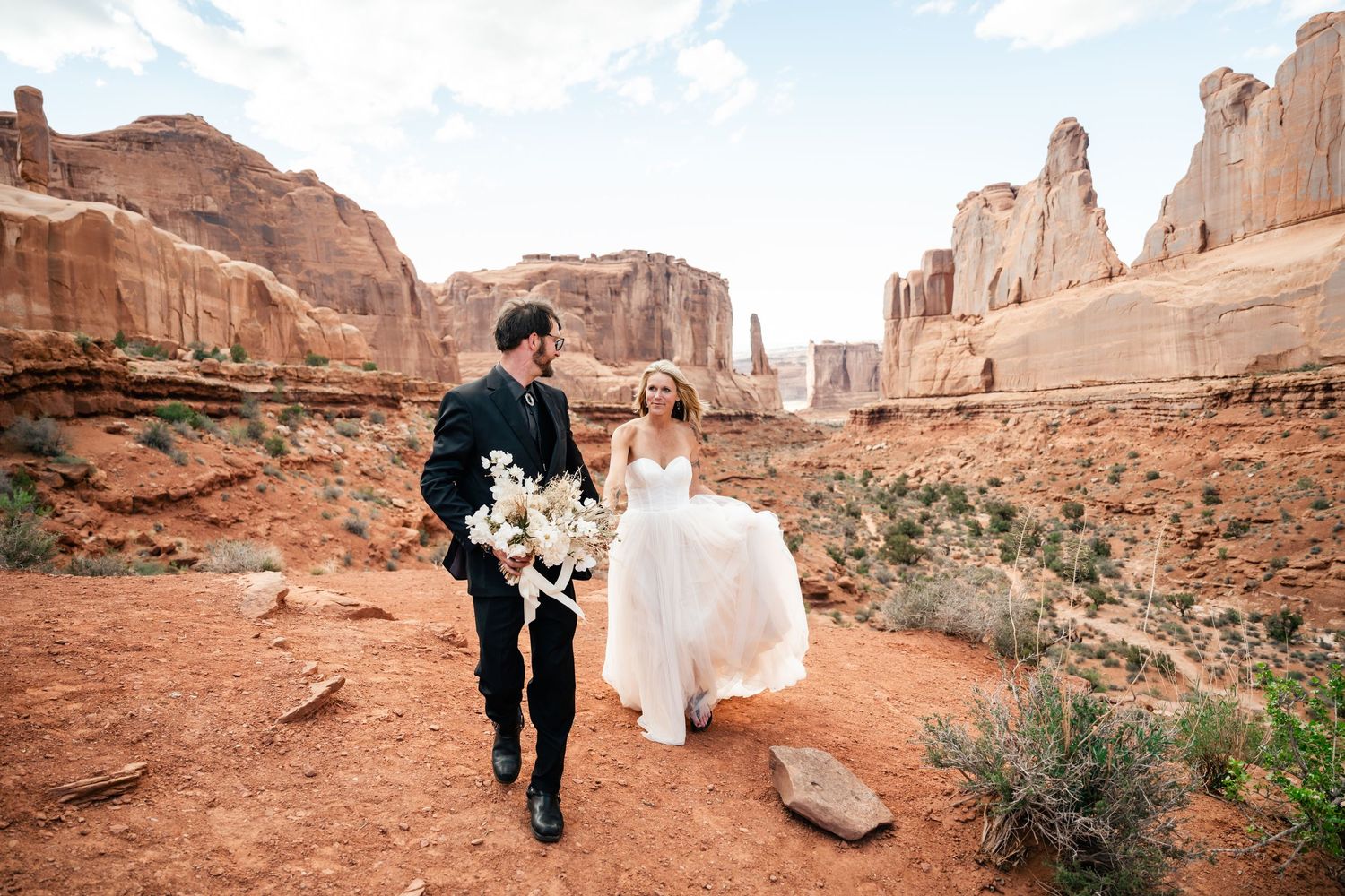 A couple in wedding attire walks through the stunning red rock formations of Arches National Park in Utah.