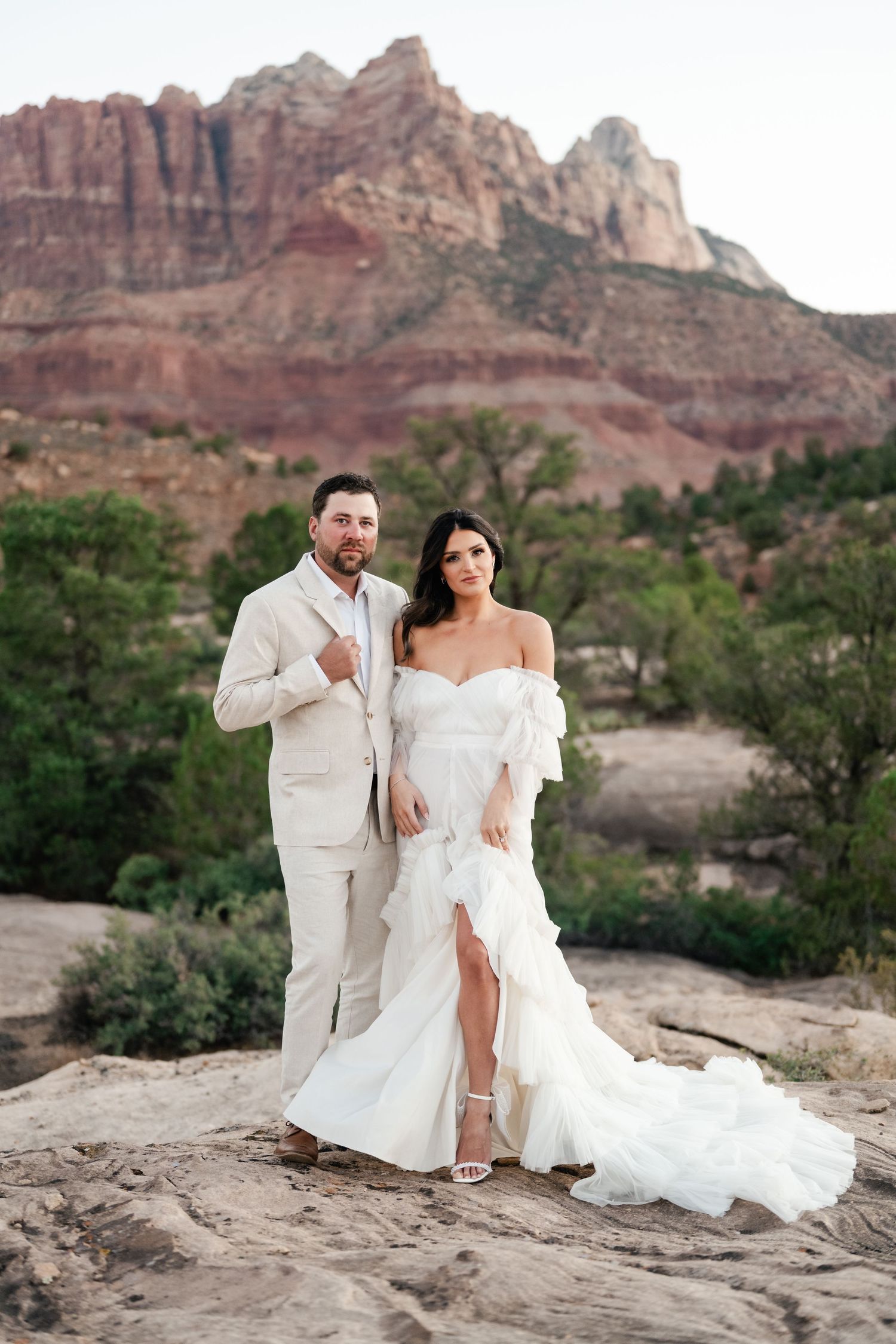 Zion destination photographer. Wedding couple poses against stunning red rock formations in a desert landscape at sunset.