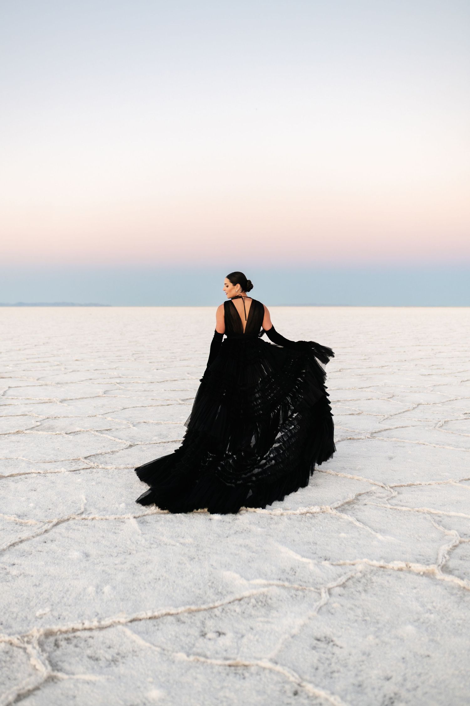 Utah Salt Flats Photographer. A figure in a flowing black gown poses dramatically on white salt flats during sunset with pastel sky above.