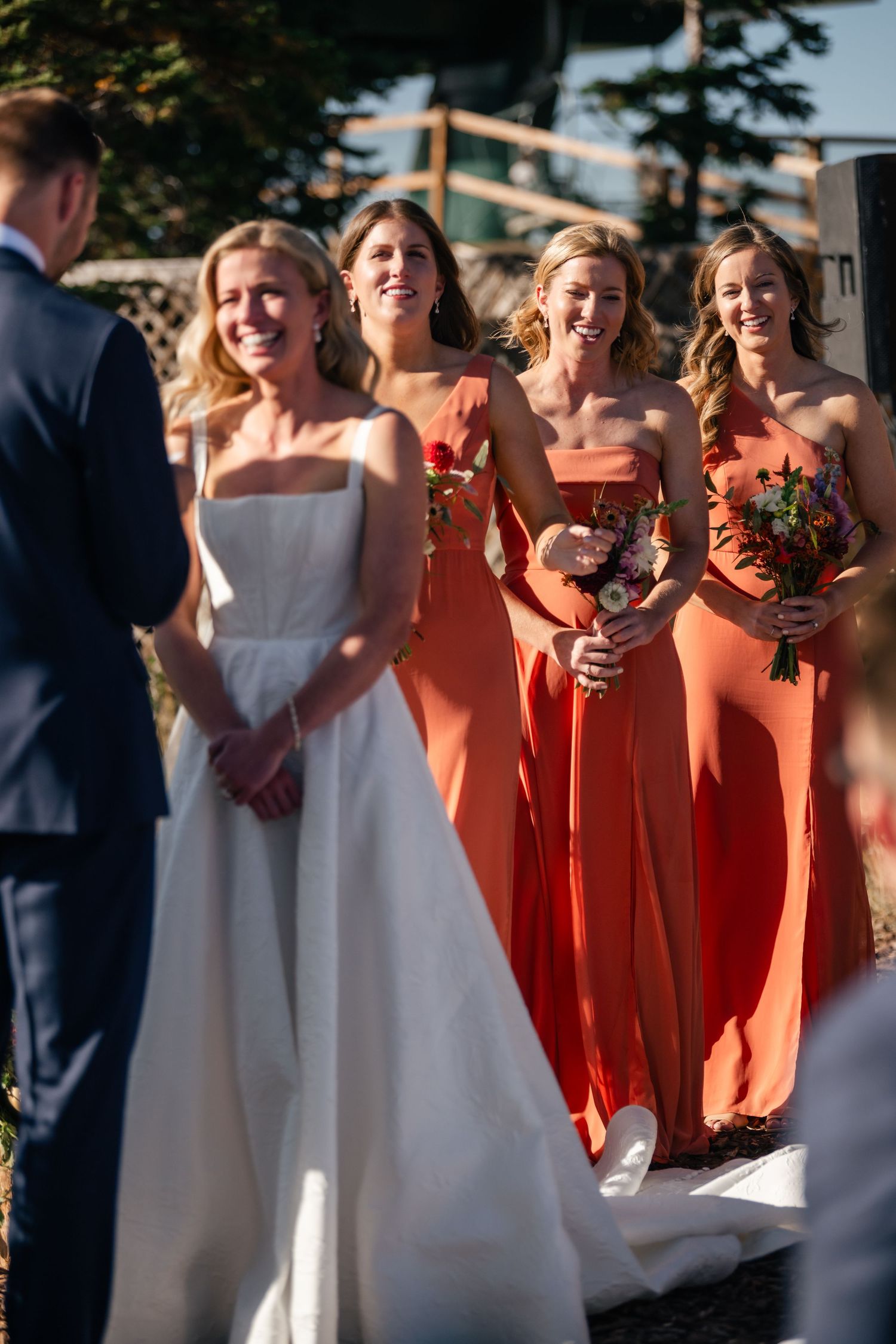 A wedding party stands outdoors with bridesmaids wearing coral-colored dresses alongside a bride in a classic white gown.