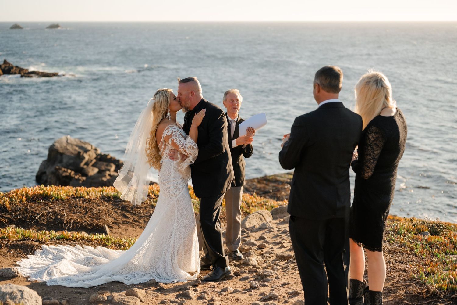 Small wedding group gathers on a dramatic coastal bluff during golden hour.