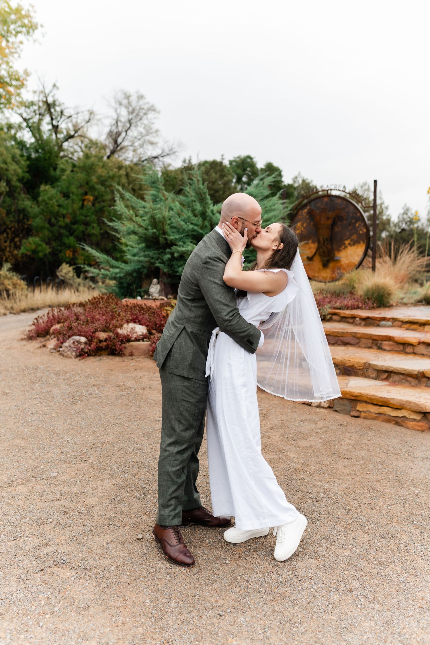 A couple in wedding attire embraces on a rustic outdoor path surrounded by autumn foliage.