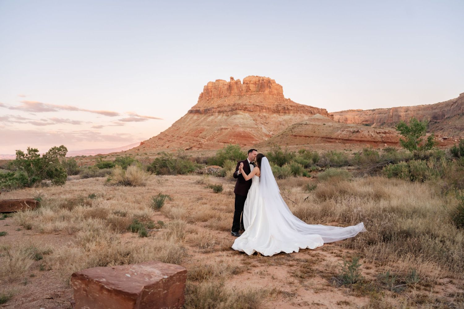 Bride and groom share a kiss before a stunning red rock butte in desert at dusk.