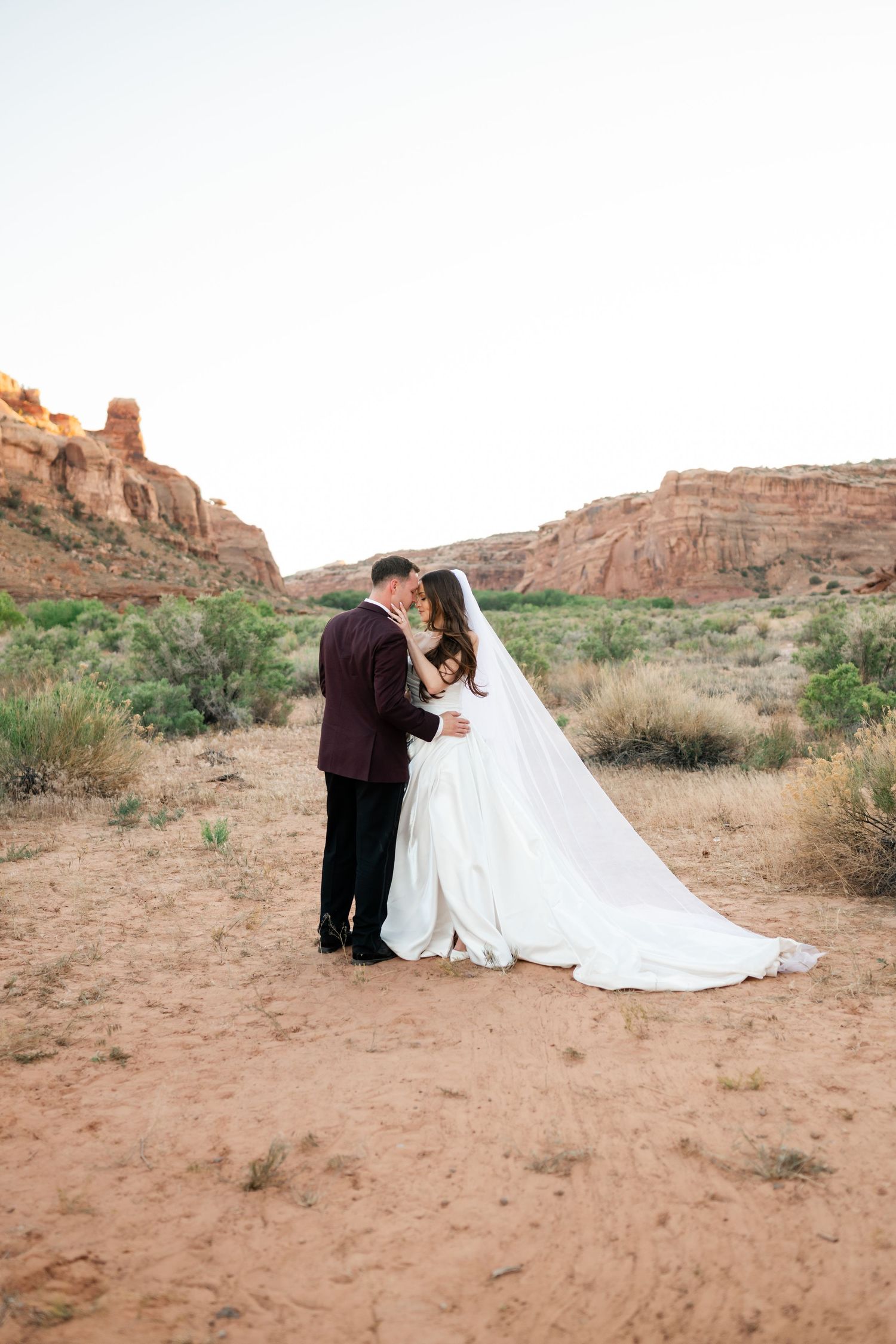 Bride and groom share intimate moment in red rock desert, her cathedral veil trails behind as they embrace at golden hour.