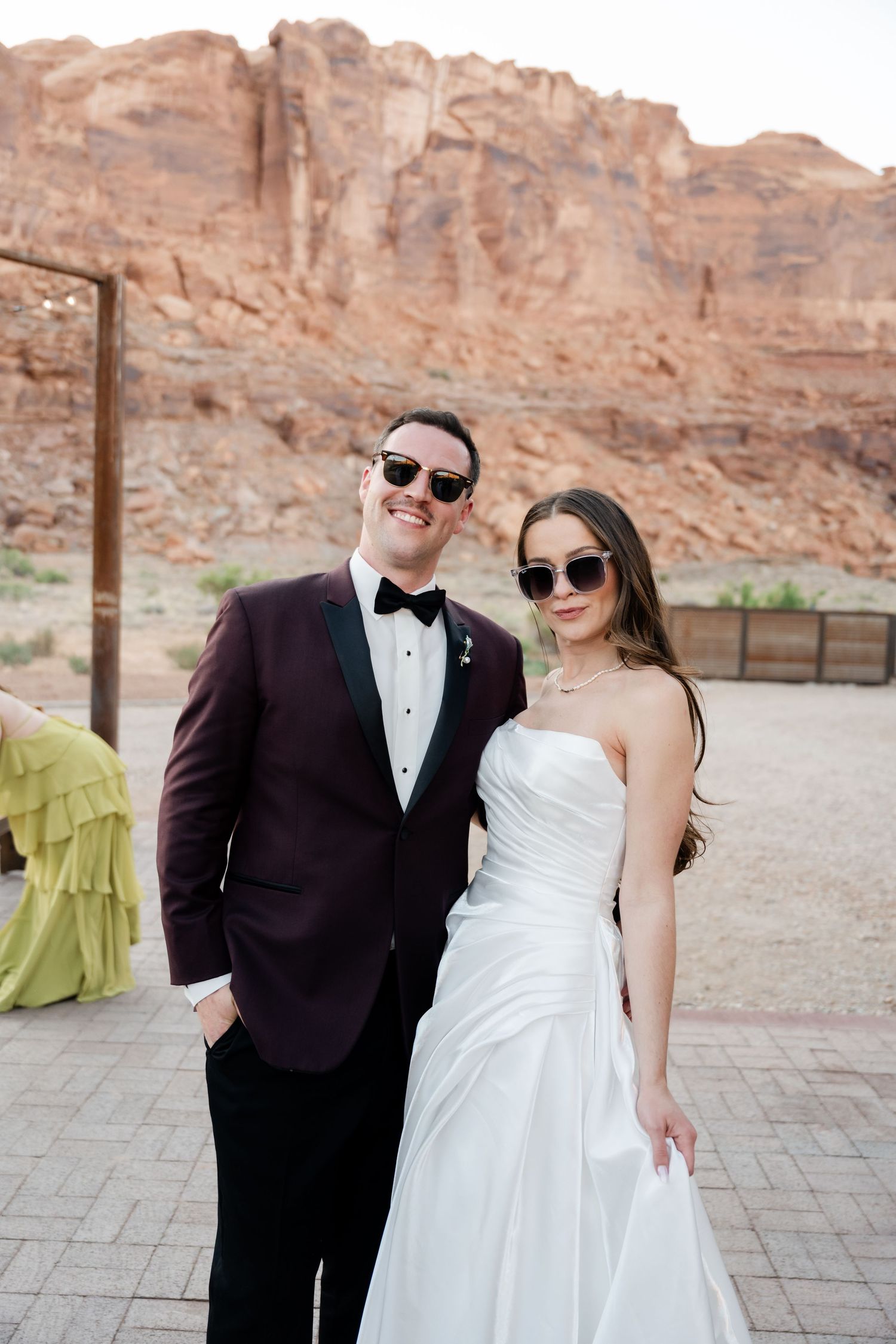 Bride and groom in sunglasses pose together at outdoor desert wedding with red rock formations in background.