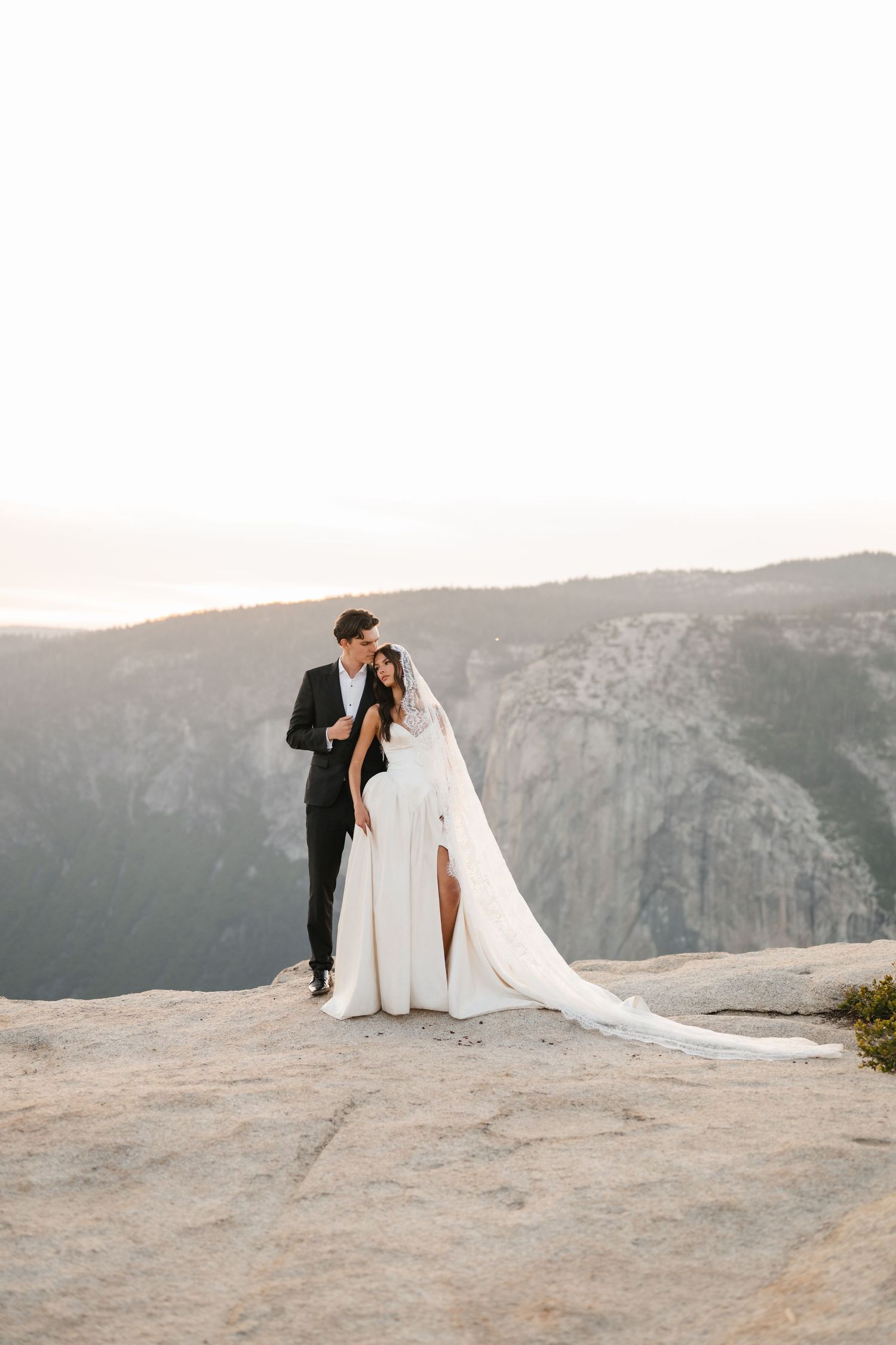 Couple embraces in wedding attire on rocky mountain overlook at golden hour.