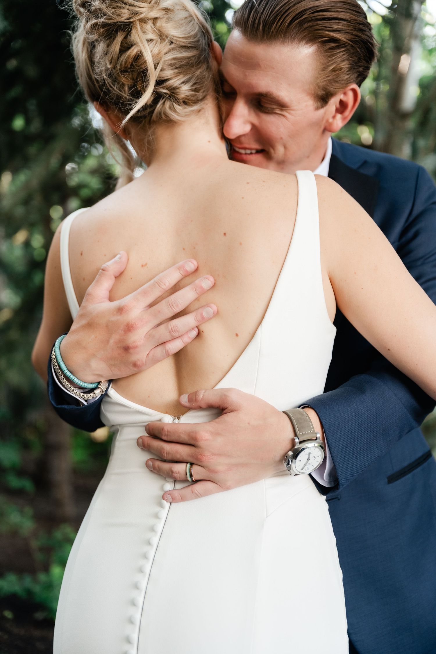 Groom in navy suit embracing bride in backless white wedding gown while smiling outdoors in garden of the Washington School House Hotel.