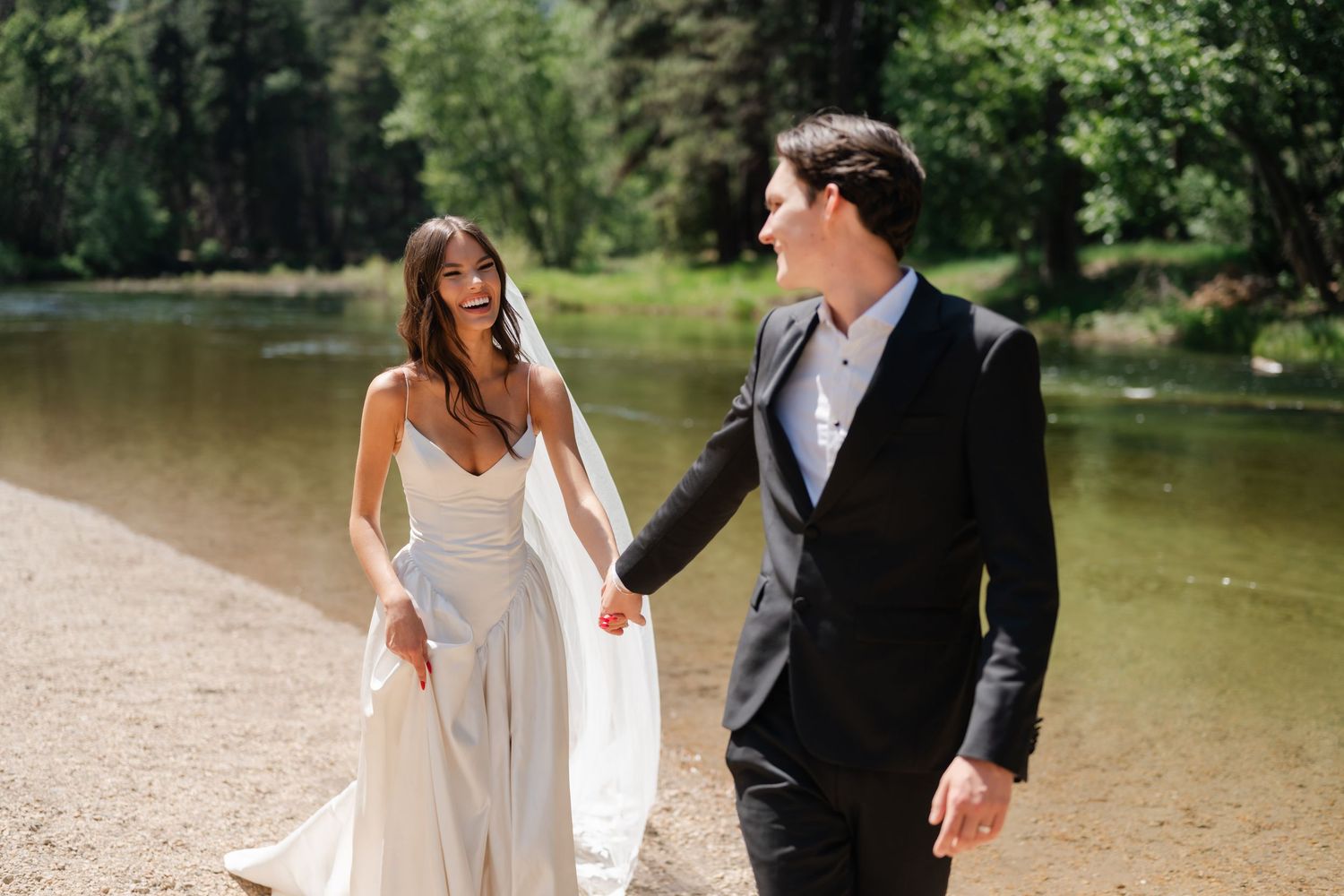Bride in white gown and groom in black suit walk hand in hand near a serene river surrounded by lush green trees.