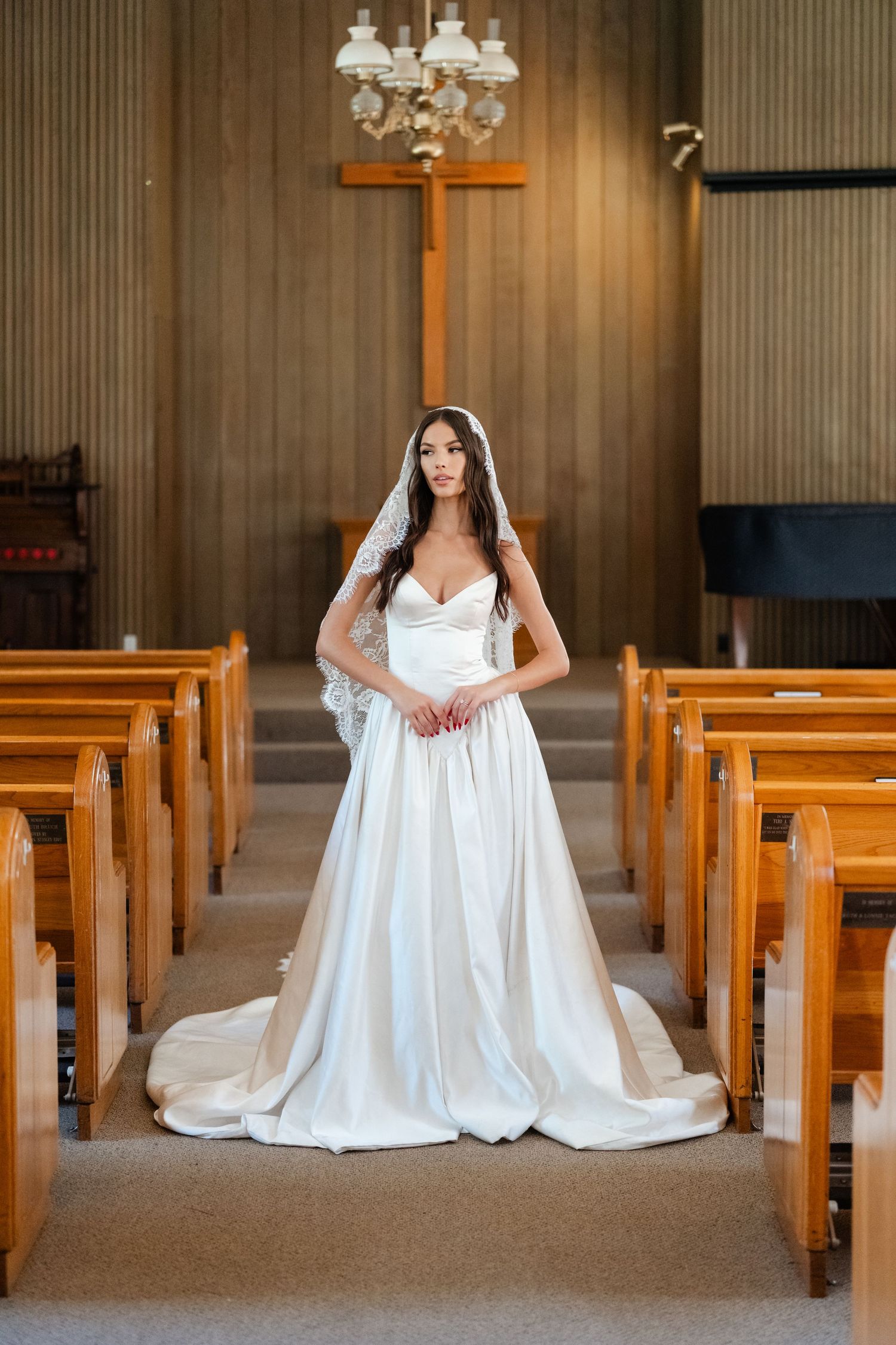 Elegant bride in strapless satin ball gown stands in church aisle with wooden pews and cross.