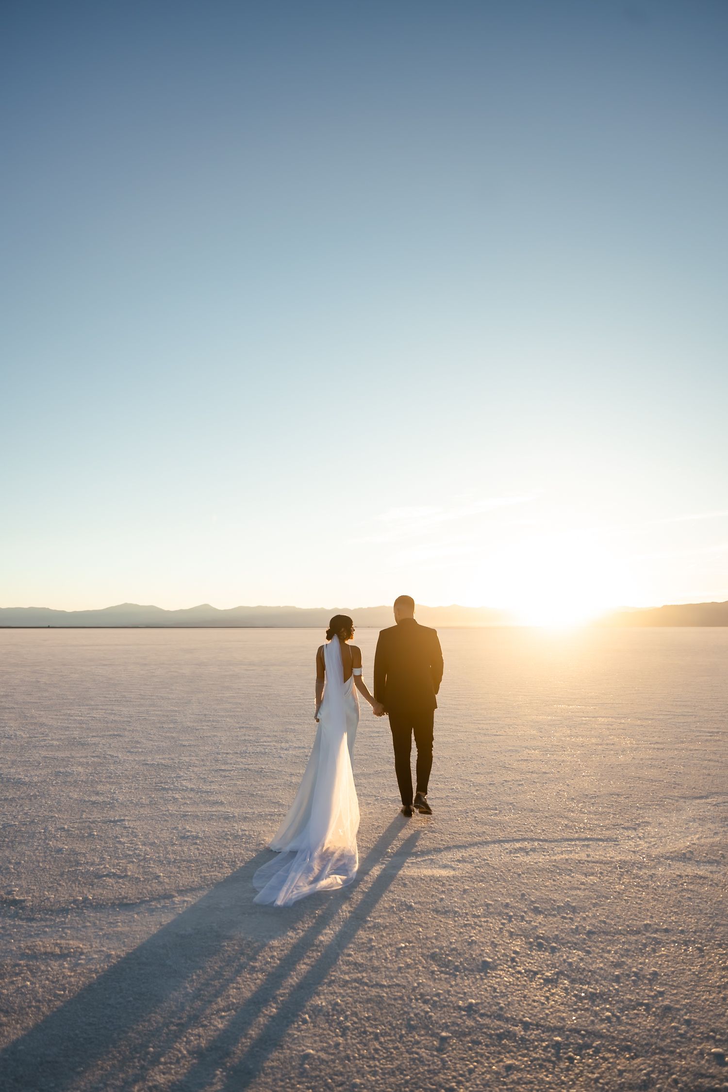 Silhouetted wedding couple walking on salt flats at sunset with long shadows cast on white ground.