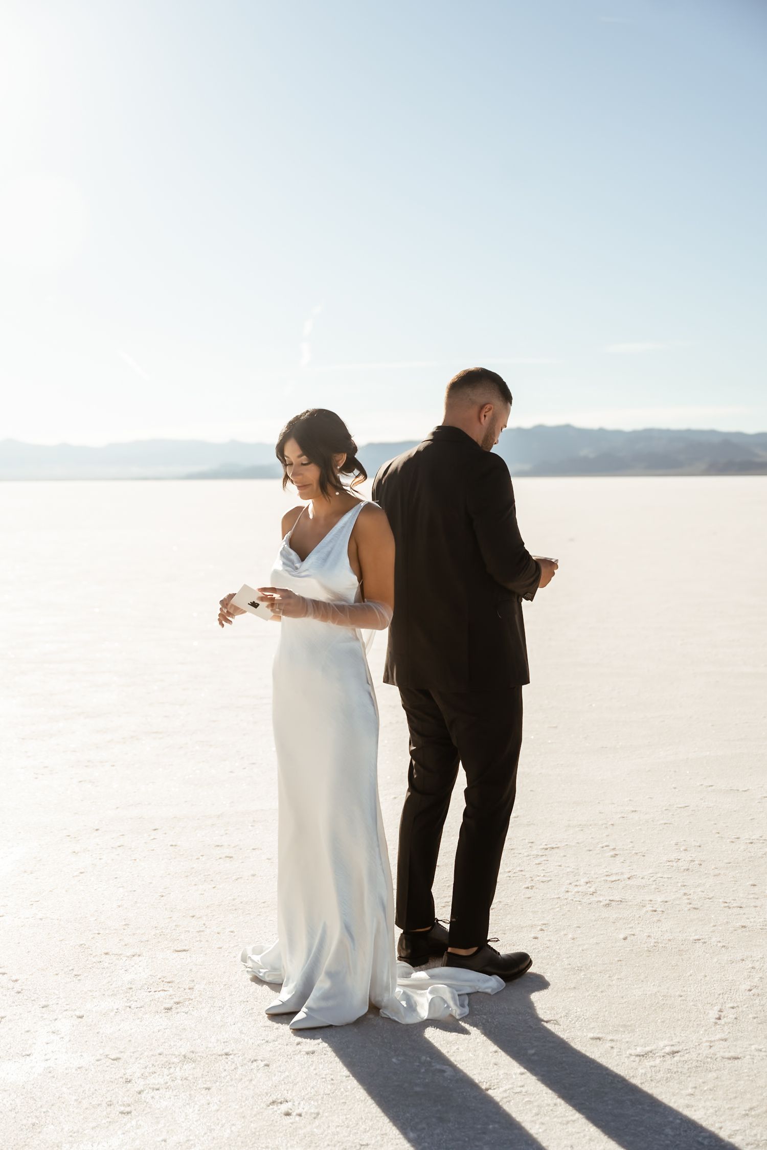 A couple stands back to back on a pristine white salt flat during their wedding.