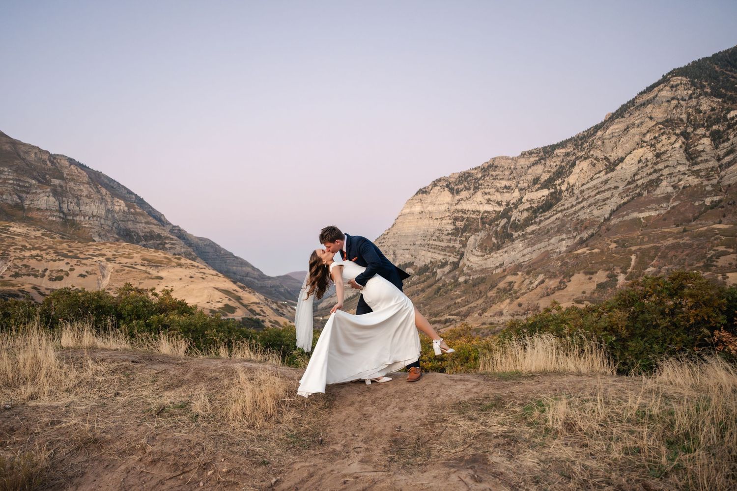 A couple in wedding attire embraces against a backdrop of rugged mountain landscapes at sunset. Utah Valley Preferred Vendor.