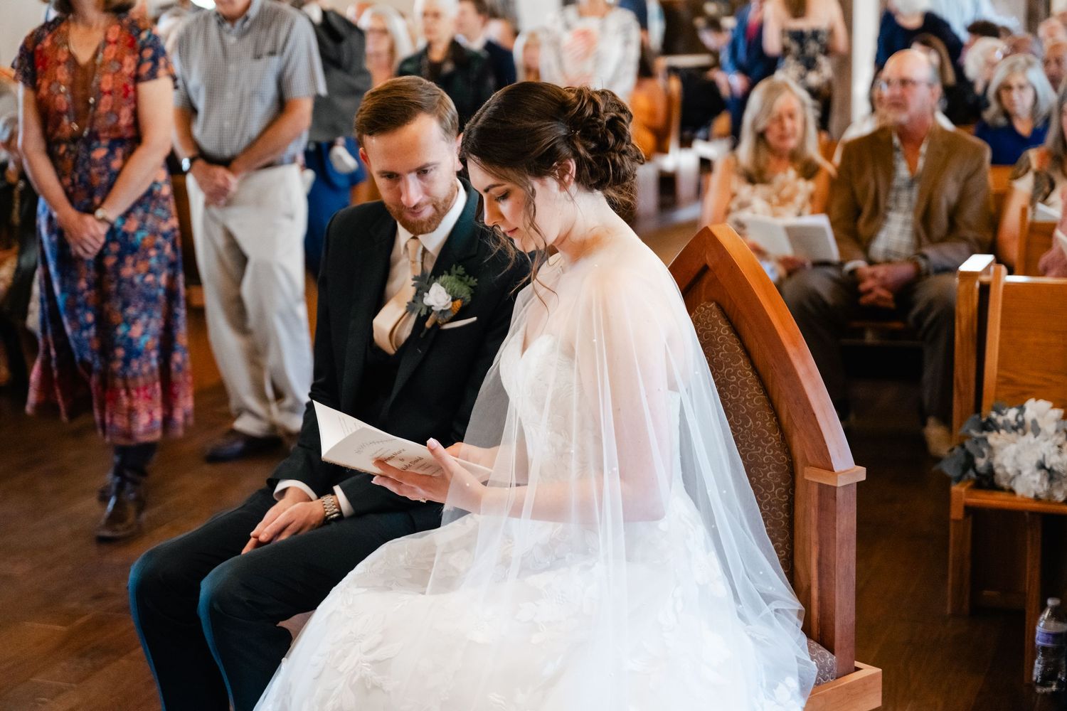 Couple seated in church pew during wedding ceremony while guests look on.