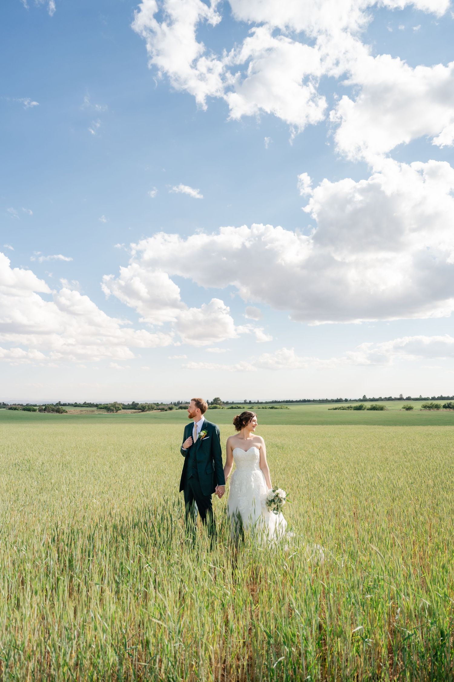 A couple walks through a vast wheat field under bright blue skies with puffy white clouds.