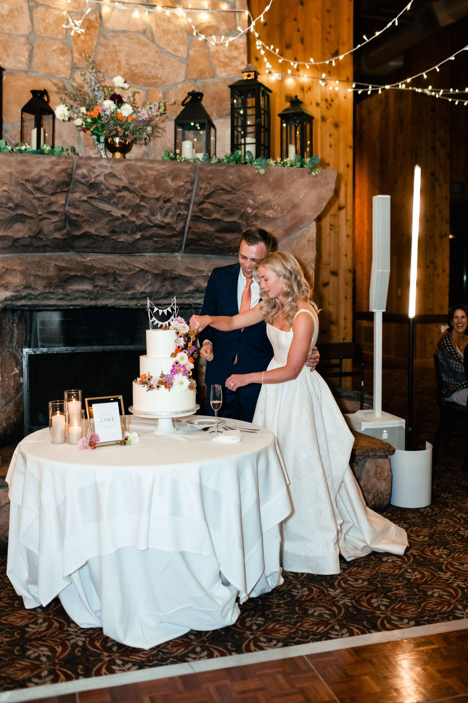 Couple cutting their white wedding cake in rustic venue with string lights.