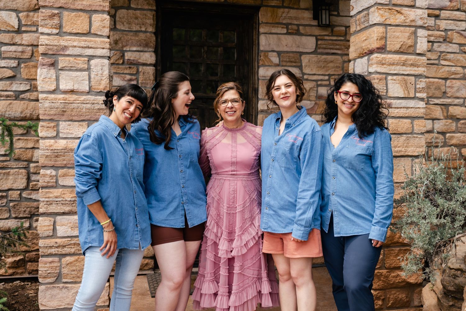A coordinated group poses in matching denim shirts and one pink dress against stone architecture.