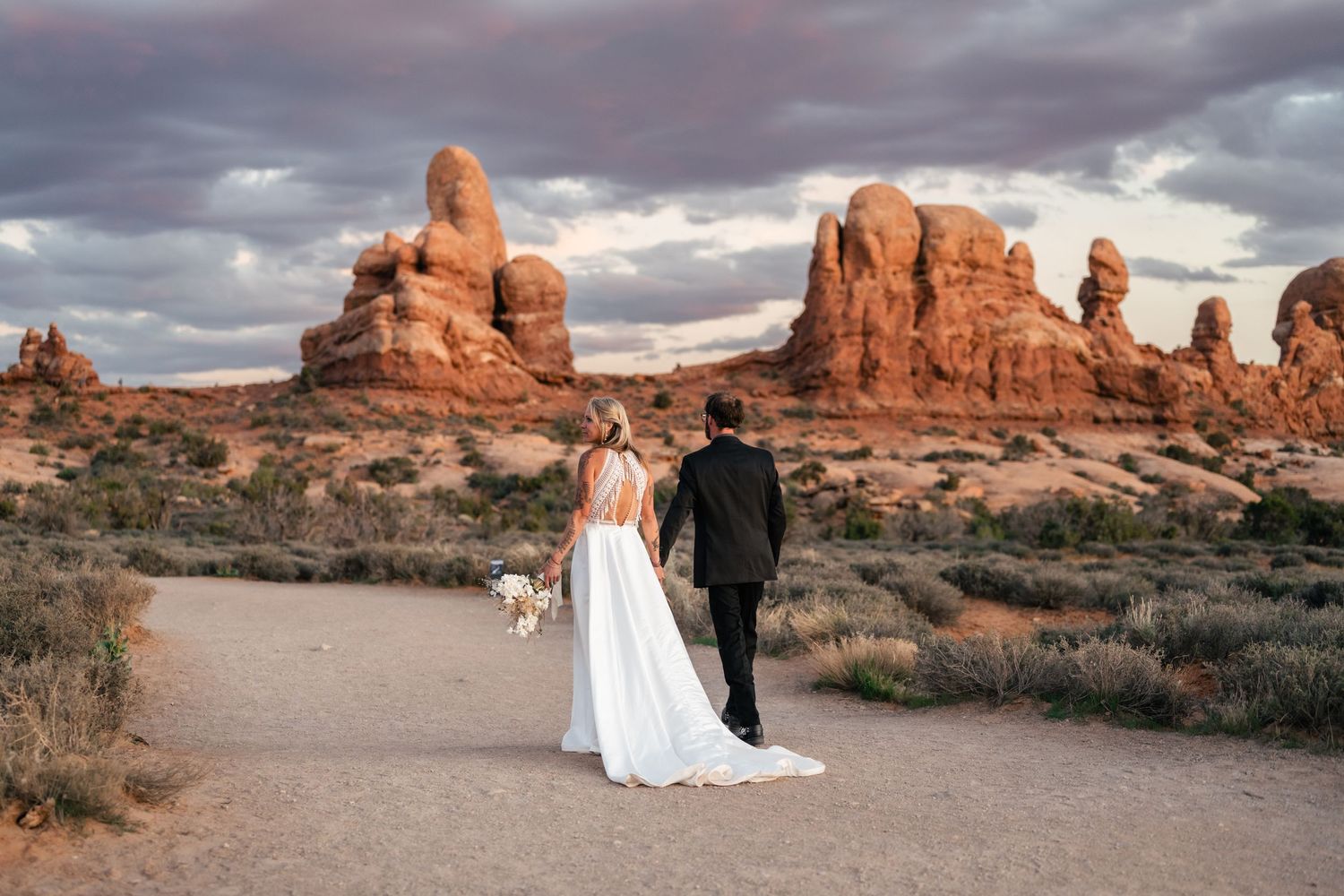 A couple in wedding attire walks through the dramatic red rock formations at Arches National Park during sunset. Moab elopement photographer