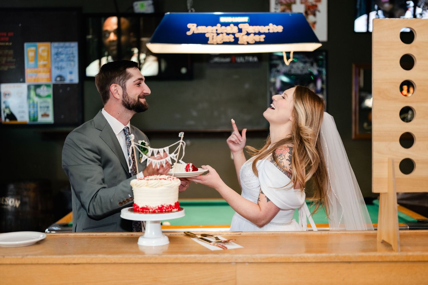 Couple sharing wedding cake at reception table with decorative lighting in background.