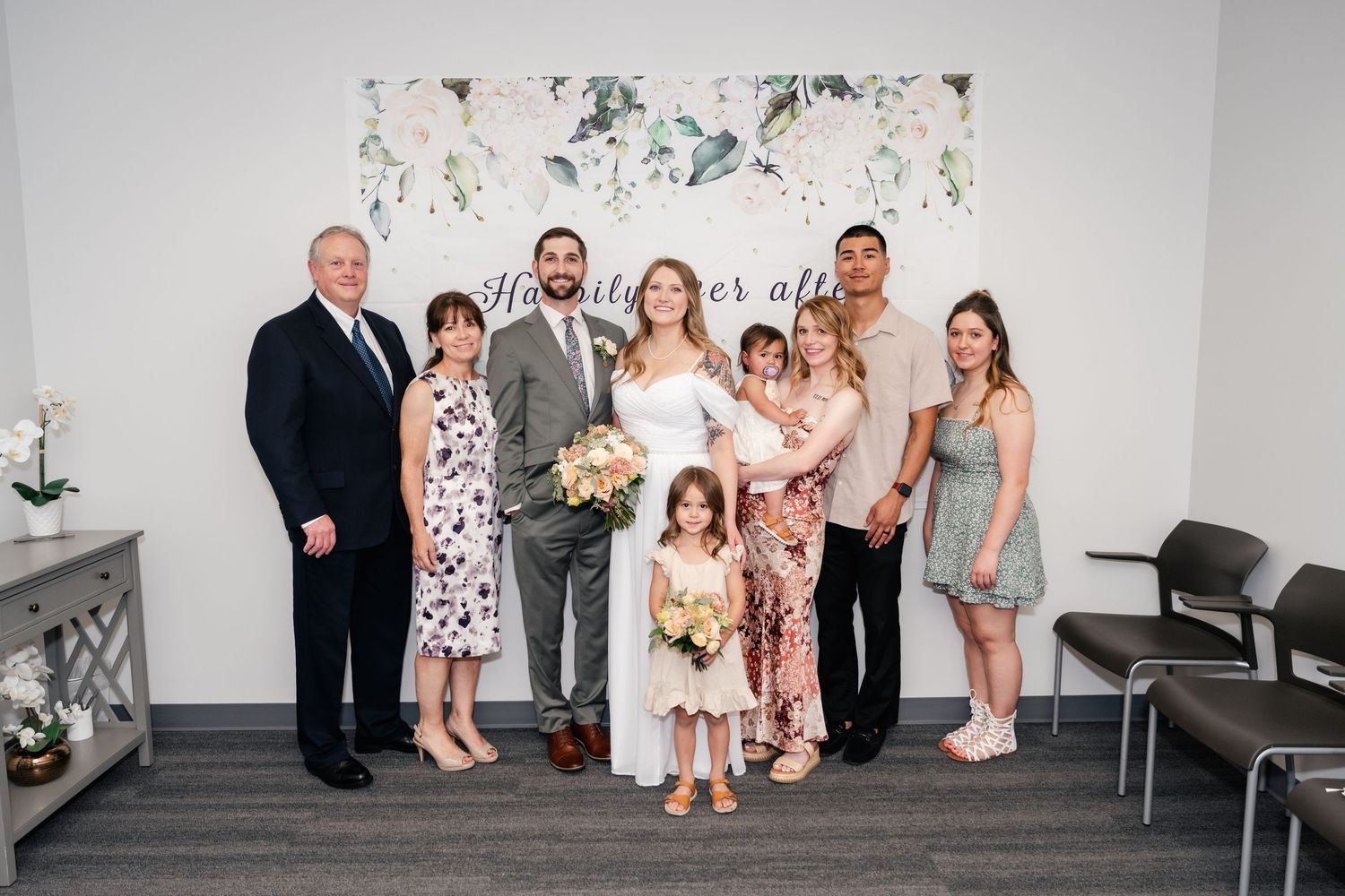 Wedding party members gather for a group photo in a bright room with floral wall decorations.