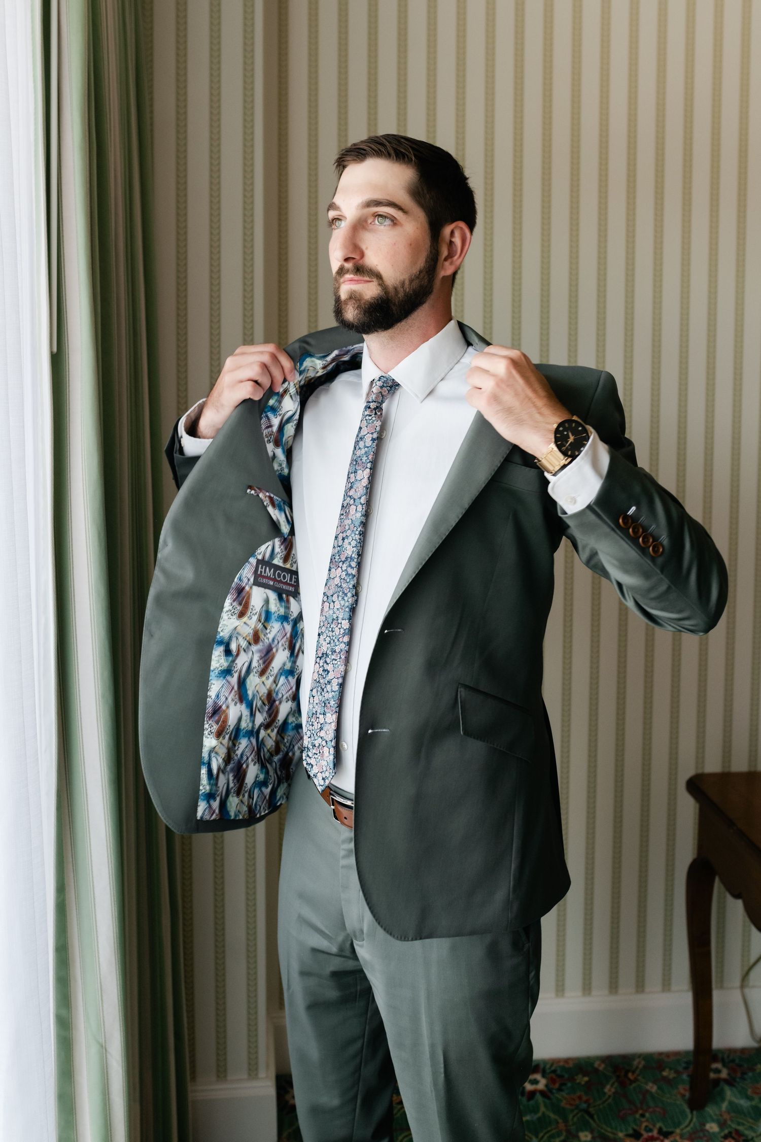 A person in a dark suit adjusts their tie while standing in a room with striped wallpaper and green curtains.
