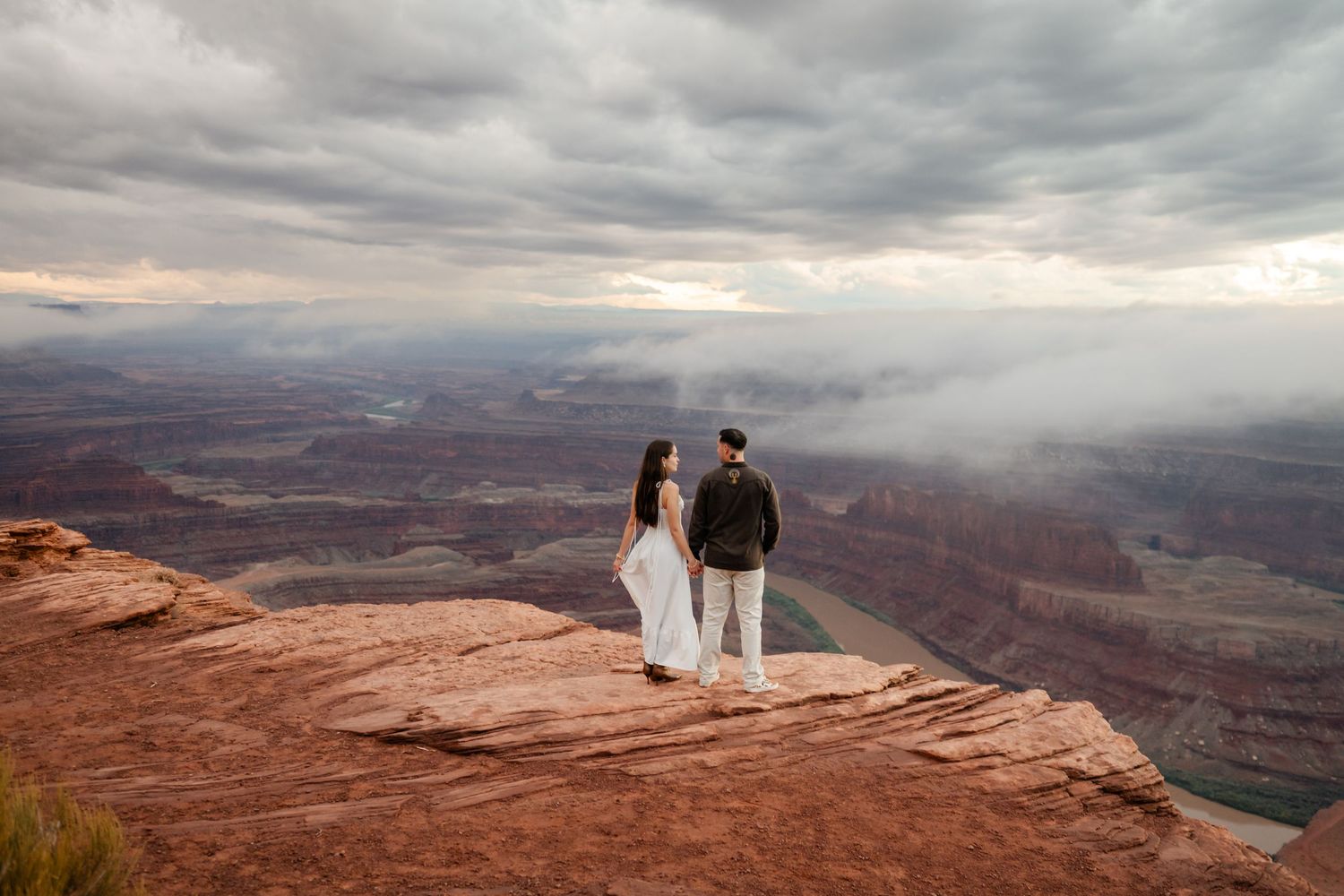 Panoramic view of a couple on a cliff edge overlooking vast desert canyons under stormy skies.