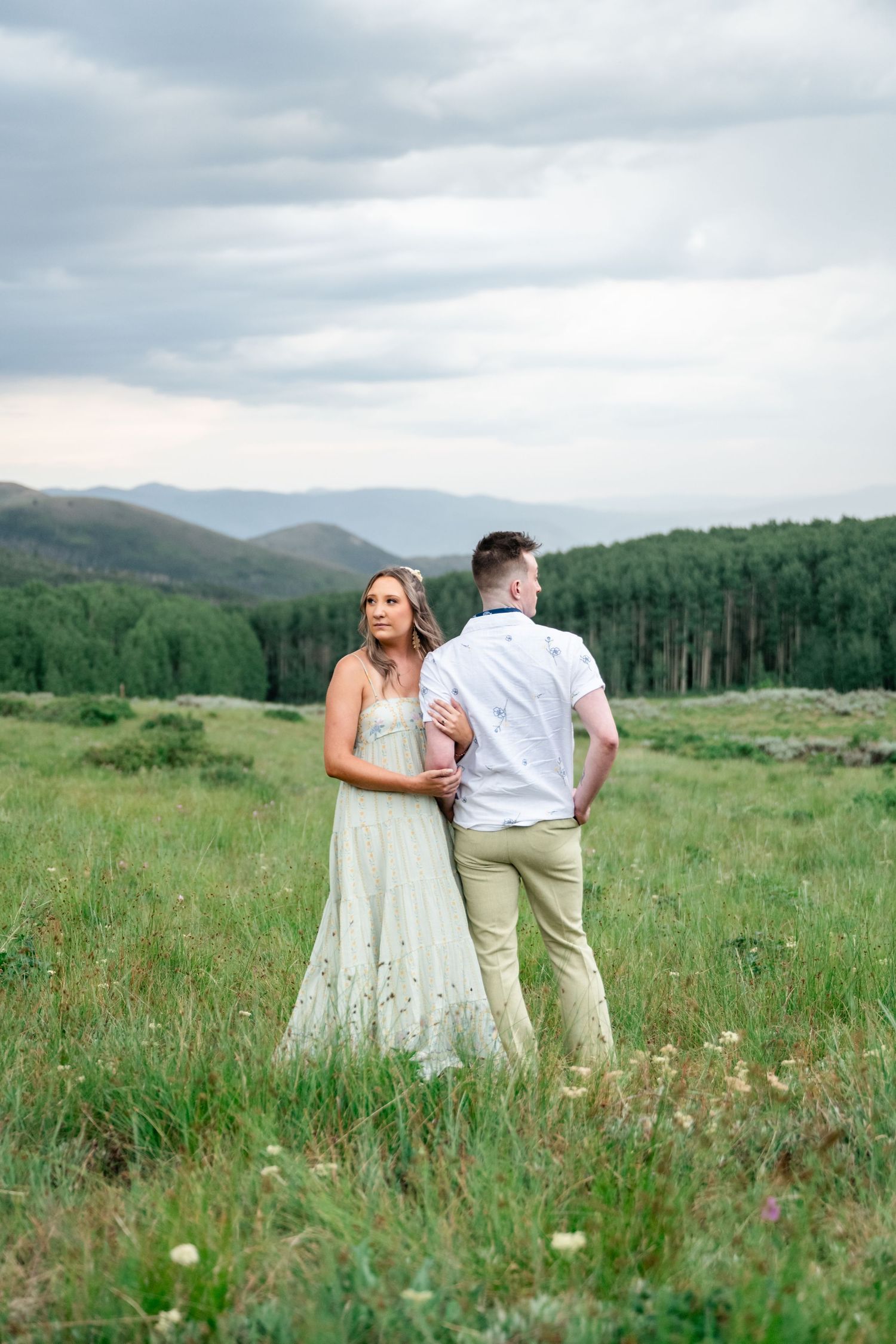 A couple in wedding attire stands together in a mountain meadow with forests and rolling hills in the background. Guardsman Pass photos.