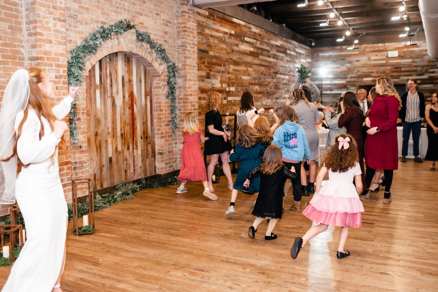 Wedding guests dancing and celebrating beneath a rustic brick archway decorated with greenery.