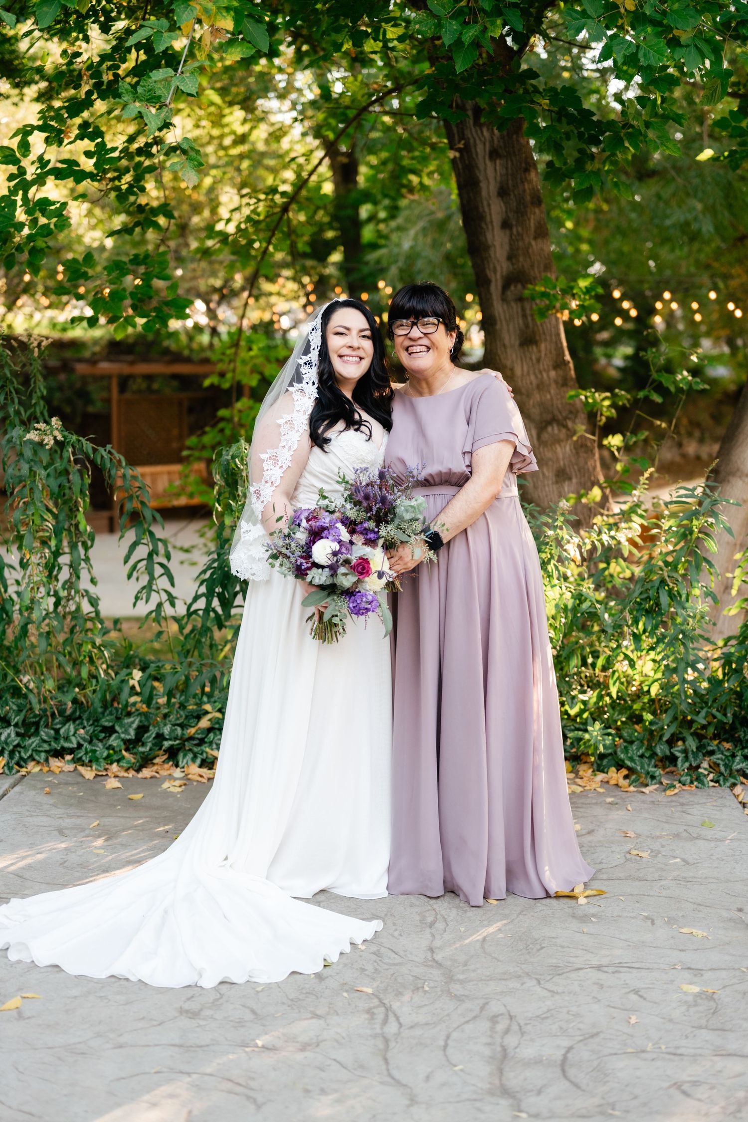 A bride in white lace gown stands with companion in mauve dress holding a purple and pink bouquet in a garden setting.