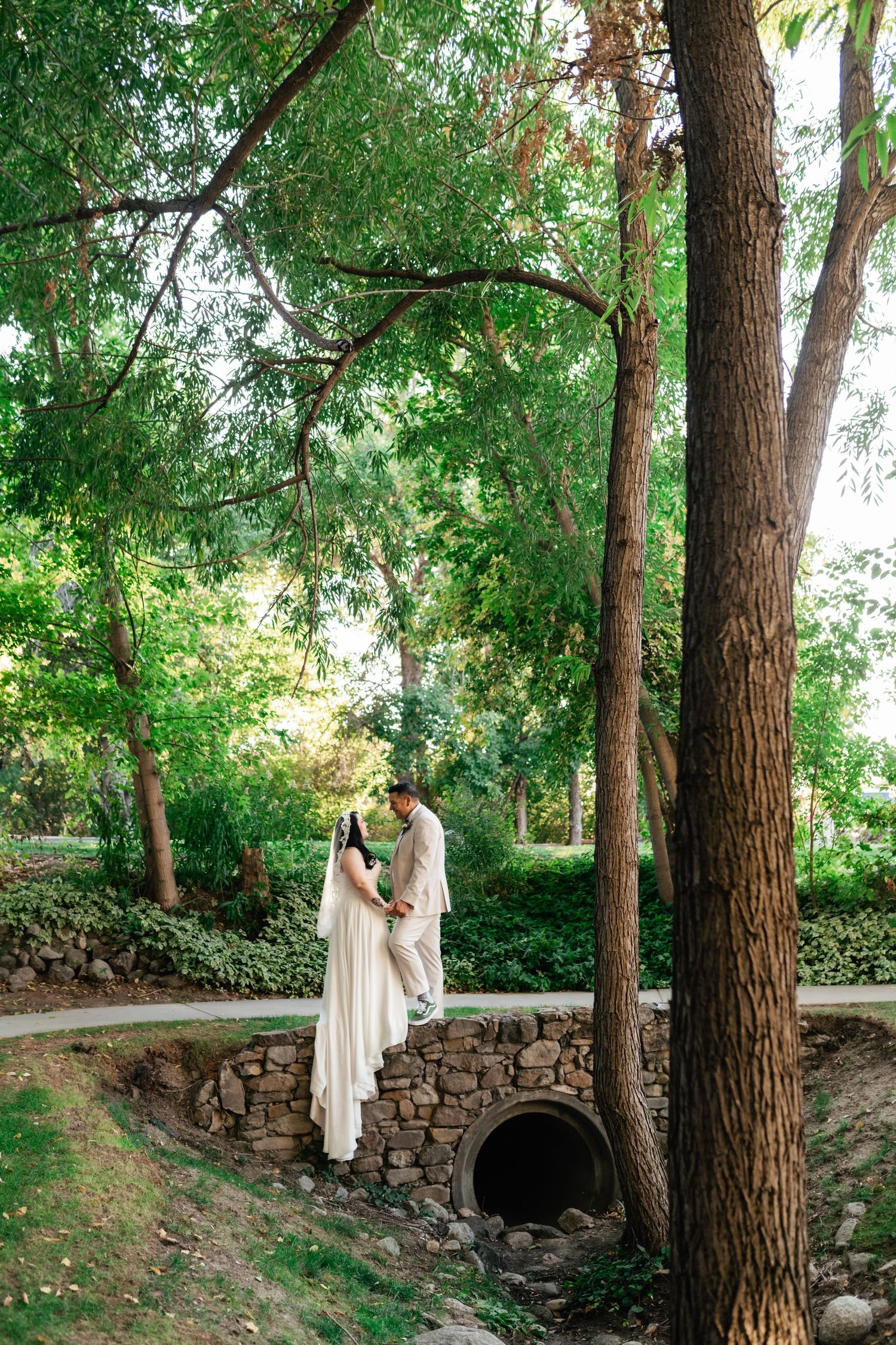 Newlyweds share an intimate moment on a rustic stone bridge surrounded by lush green trees.