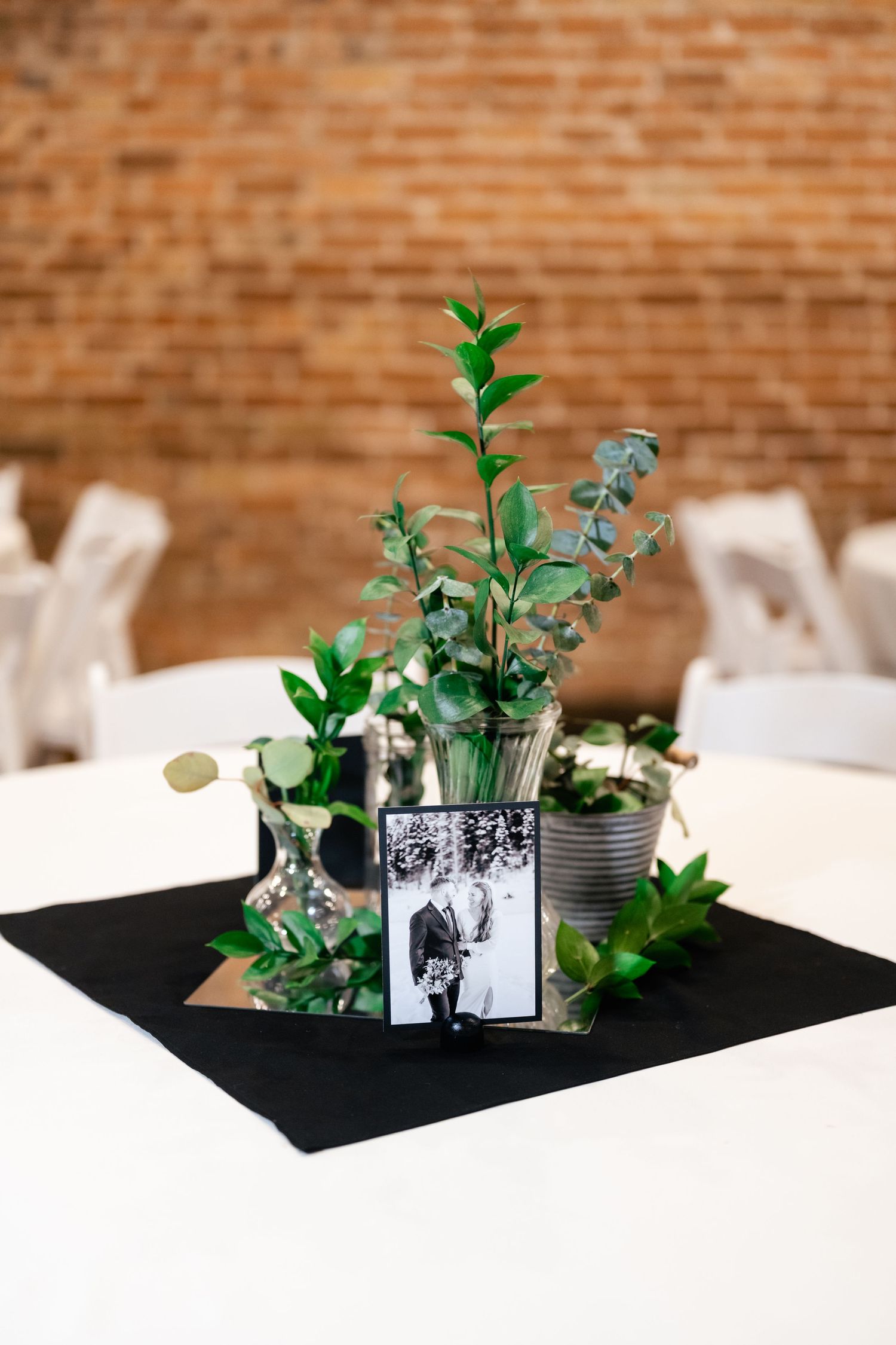 Wedding centerpiece with greenery and eucalyptus in a tin can vase with a black and white photograph on a black mat.