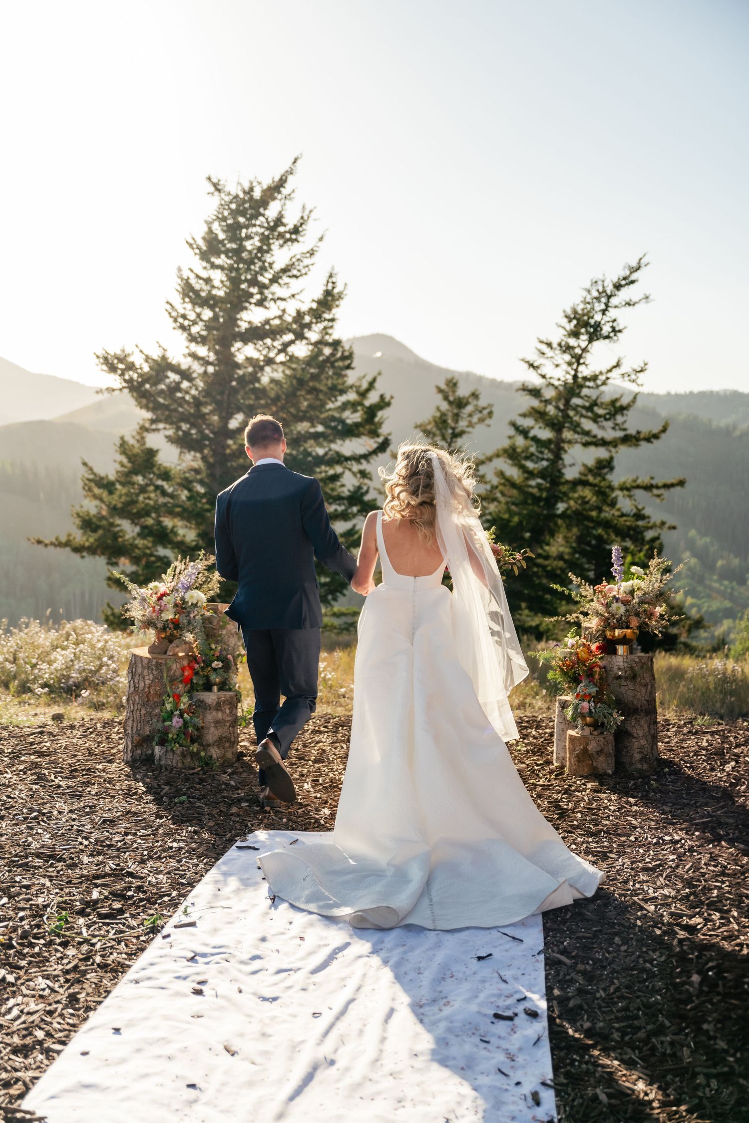 Couple walks down white aisle runner at outdoor mountain ceremony with floral arch decorations. Park City Wedding Photographer.