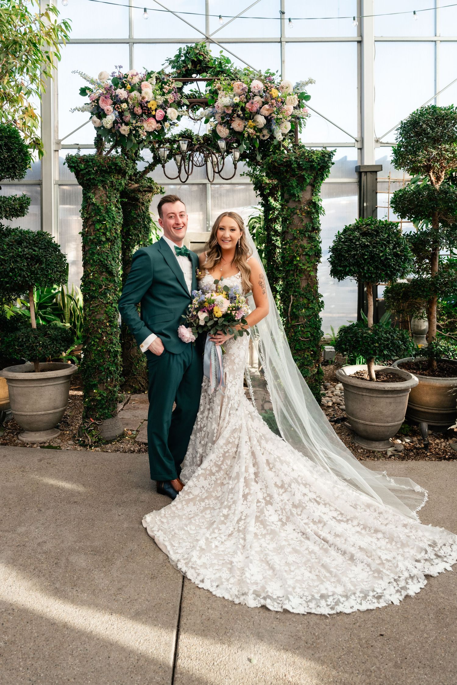 A wedding couple poses beneath an elegant floral arch with pink and white roses in a greenhouse setting with topiary trees.