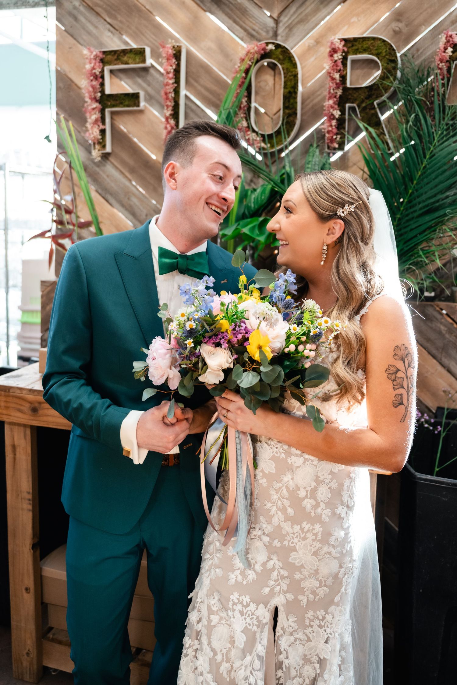 Newlyweds share a joyful moment under floral 'FOR' letters while holding colorful bouquet.