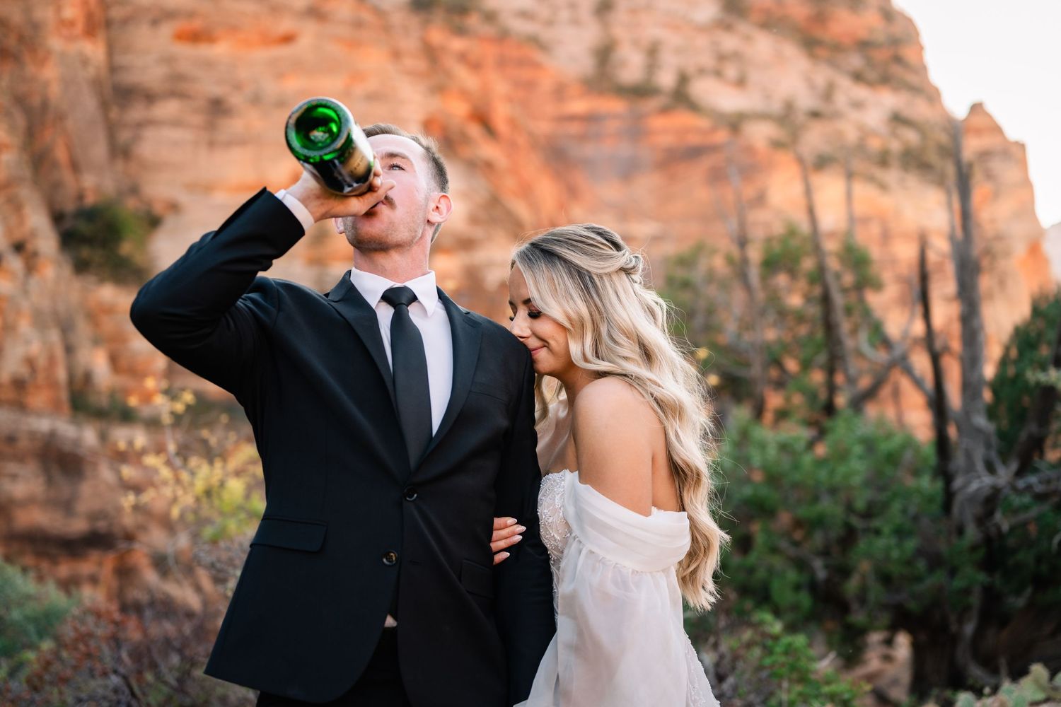 A couple in wedding attire shares a playful moment against red rock cliffs at sunset.