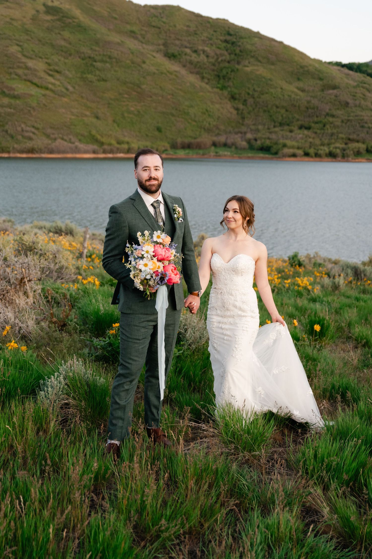 Bride and groom stand together on hillside overlooking lake with colorful spring wildflowers.