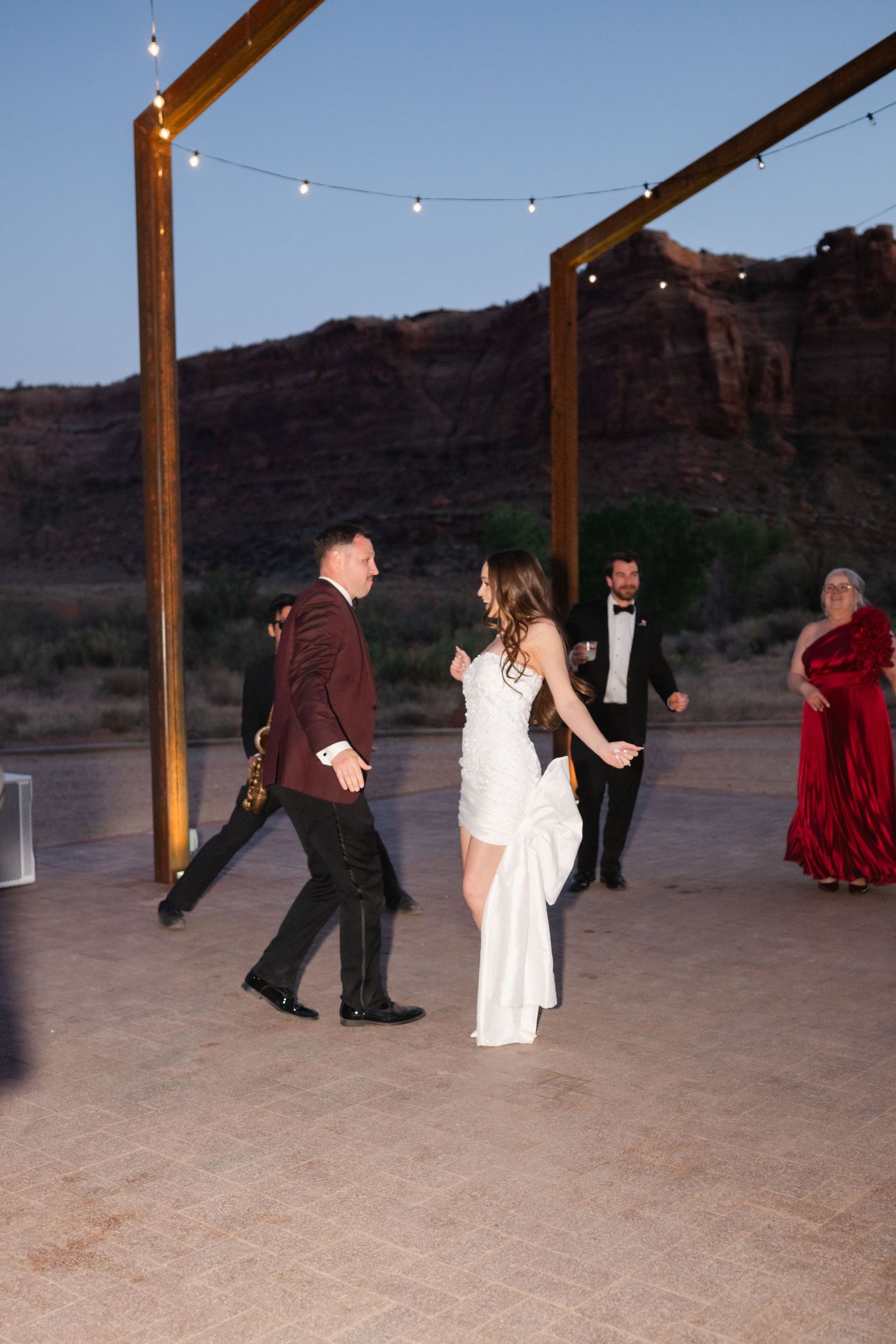 Bride and groom share first dance on outdoor patio under string lights with red rock cliffs at dusk.