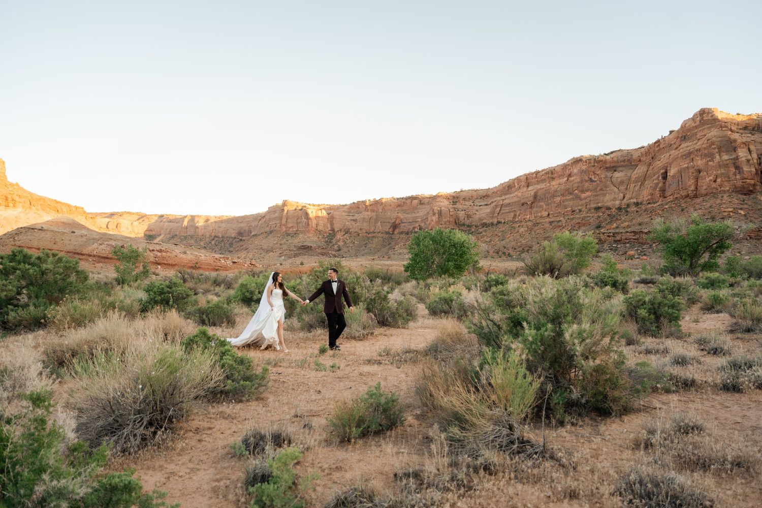 Bride and groom hold hands walking through desert scrubland with red rock canyon cliffs at golden hour.