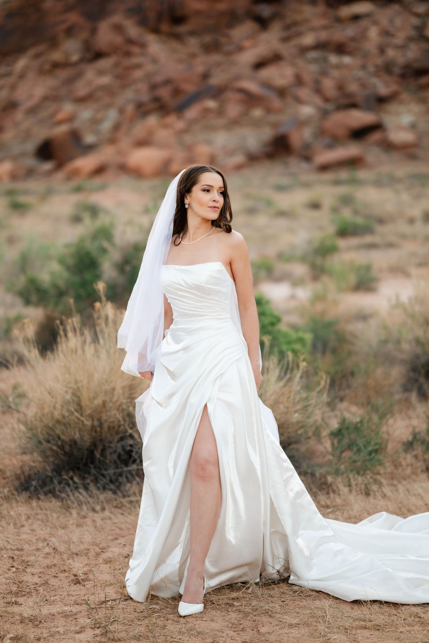 Bride in strapless white satin gown with thigh slit and veil poses in desert landscape with red rocks.