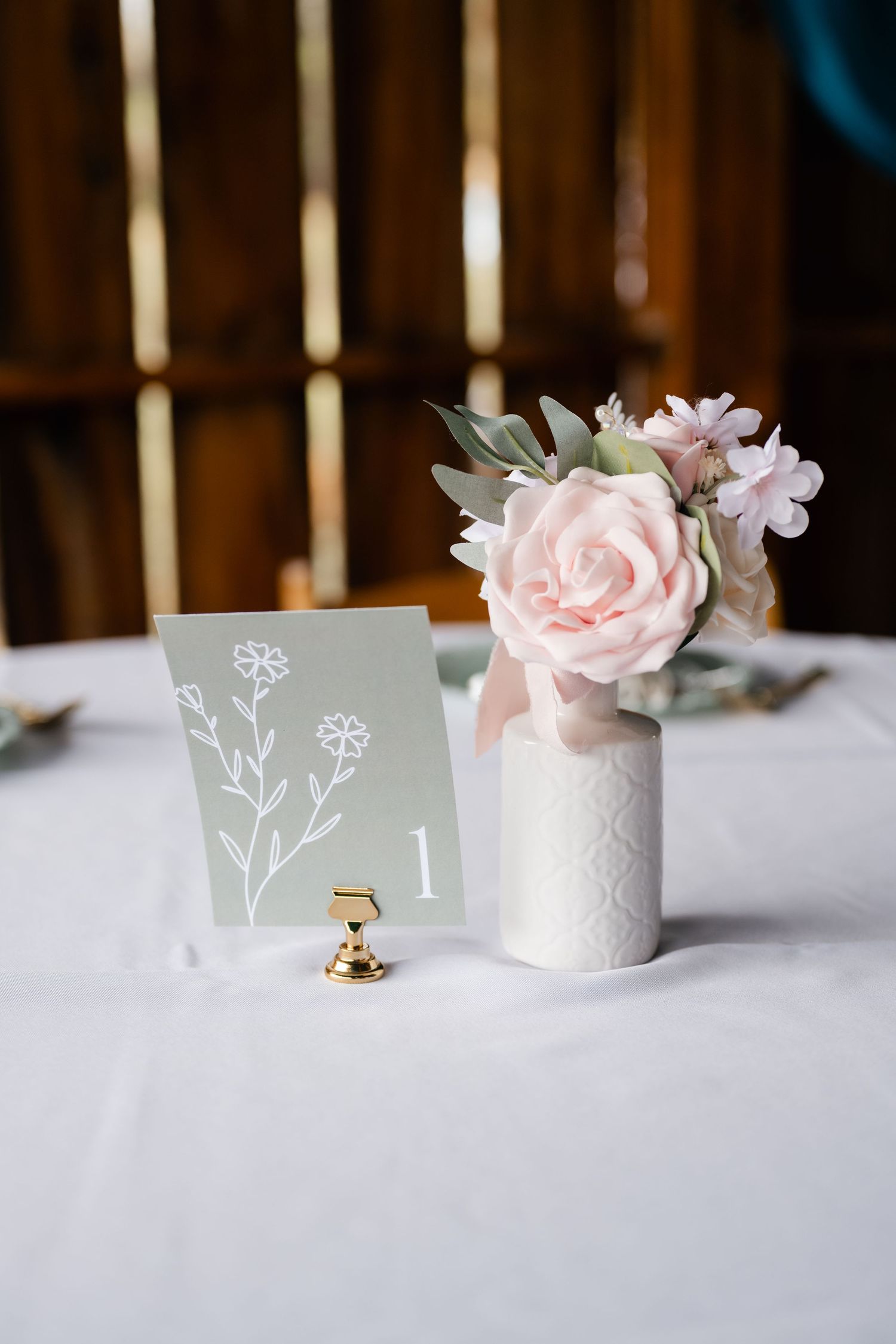 Elegant wedding table number with pink roses in white ceramic vase on white tablecloth.