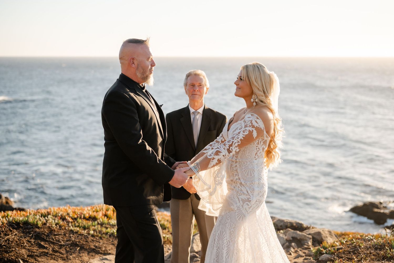 A couple exchanges vows during their seaside wedding ceremony with waves crashing against rocks in the background.