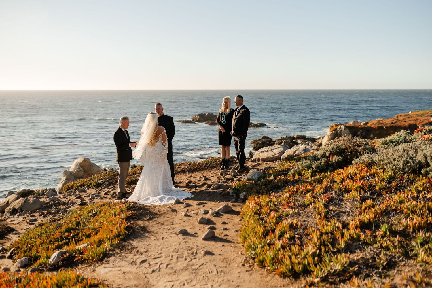 Scenic panorama of a beach wedding with attendees silhouetted against the ocean horizon.