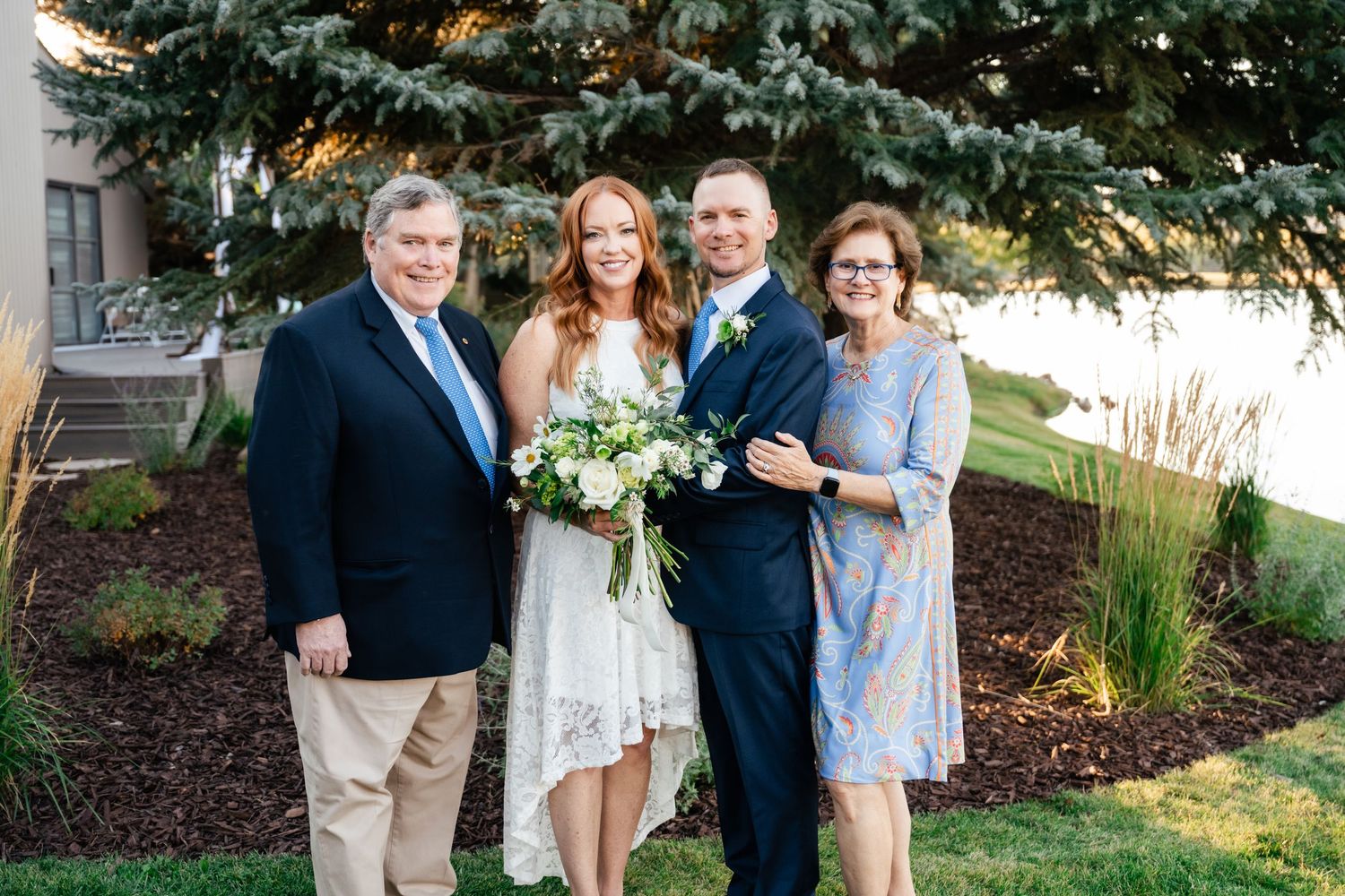 A wedding party poses in front of evergreen trees with a bride in a white dress holding a floral bouquet.