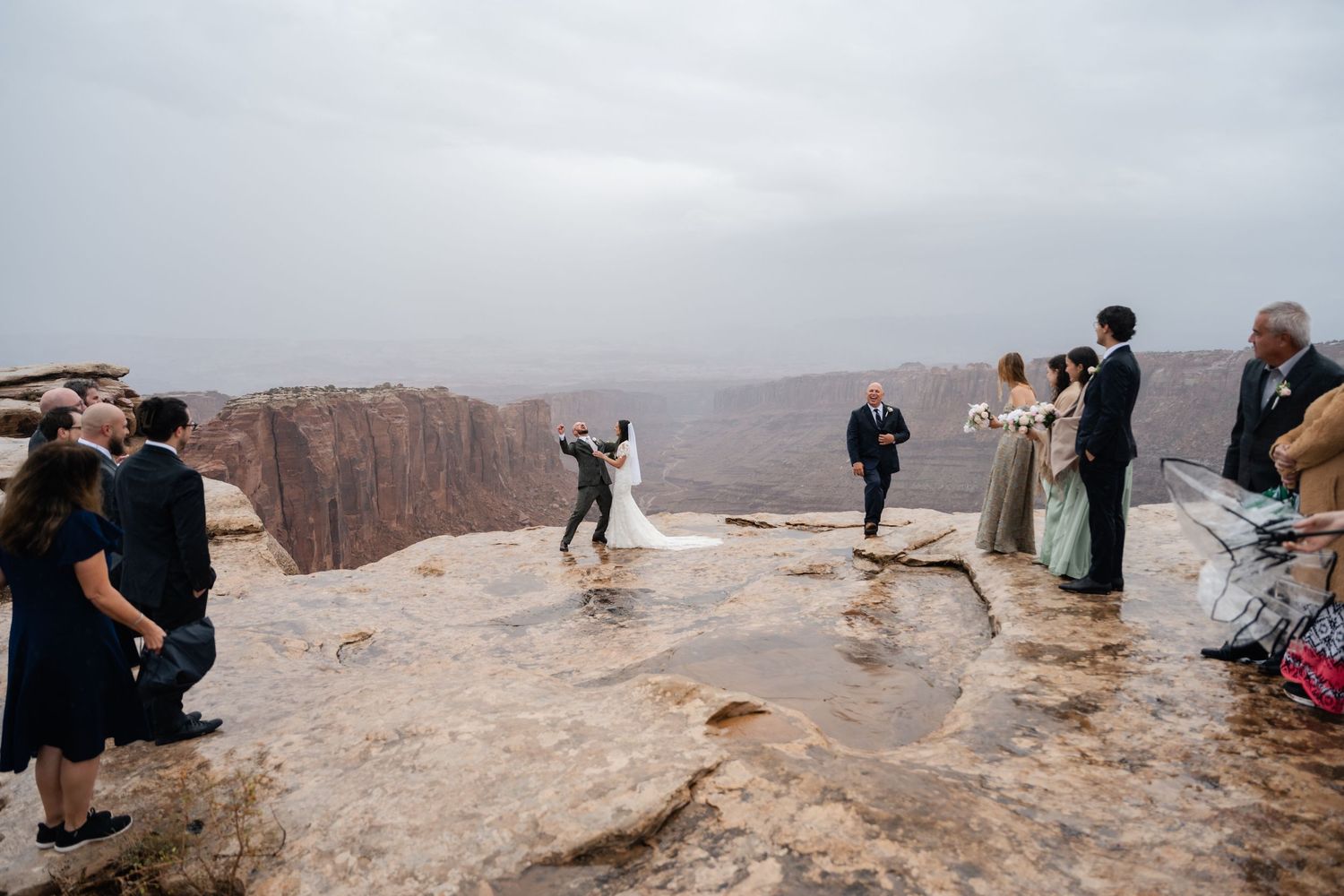 Wedding party and guests gather on scenic desert cliff for ceremony photos.