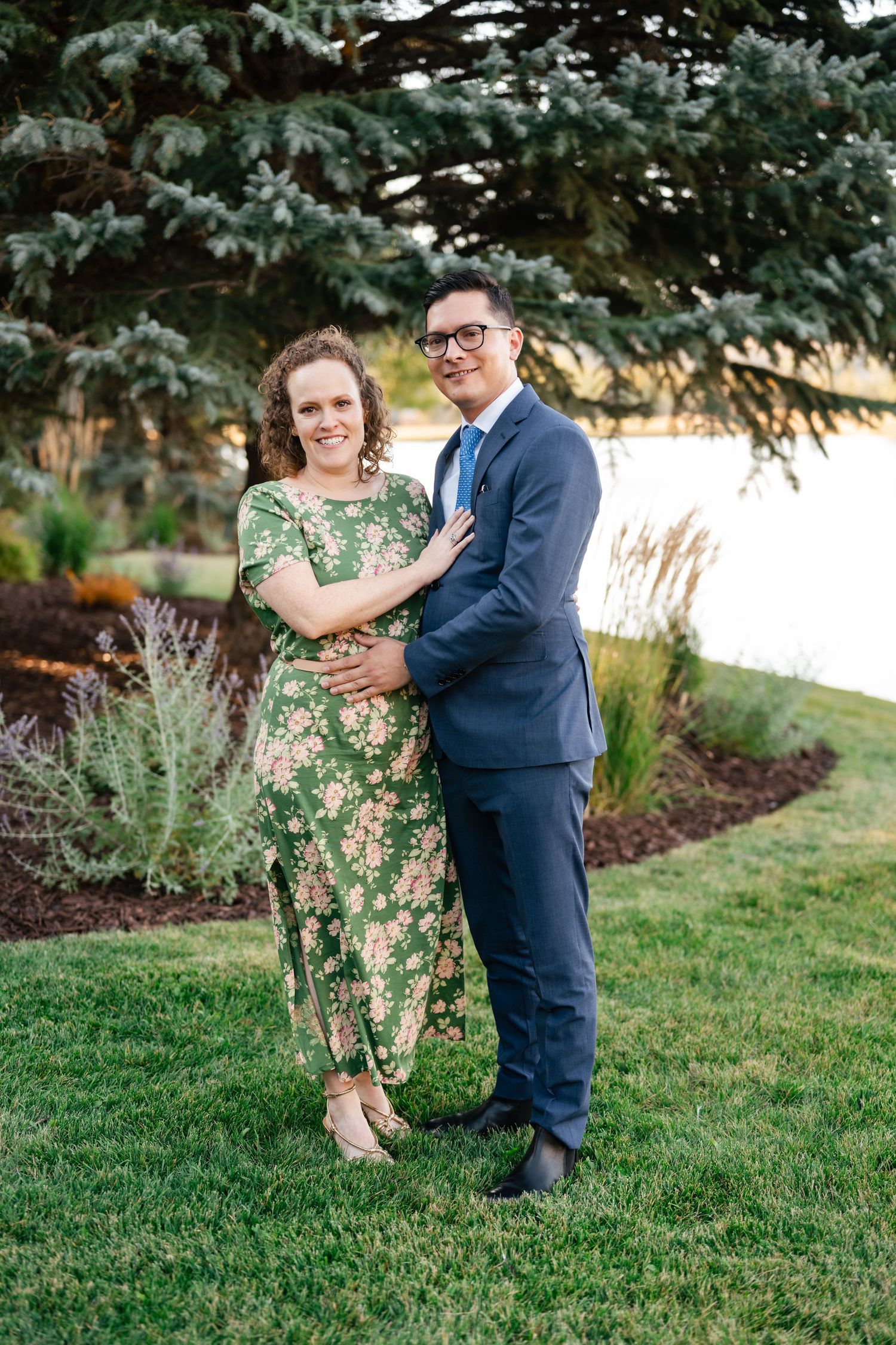 Couple poses together outdoors in front of evergreen trees during sunset.
