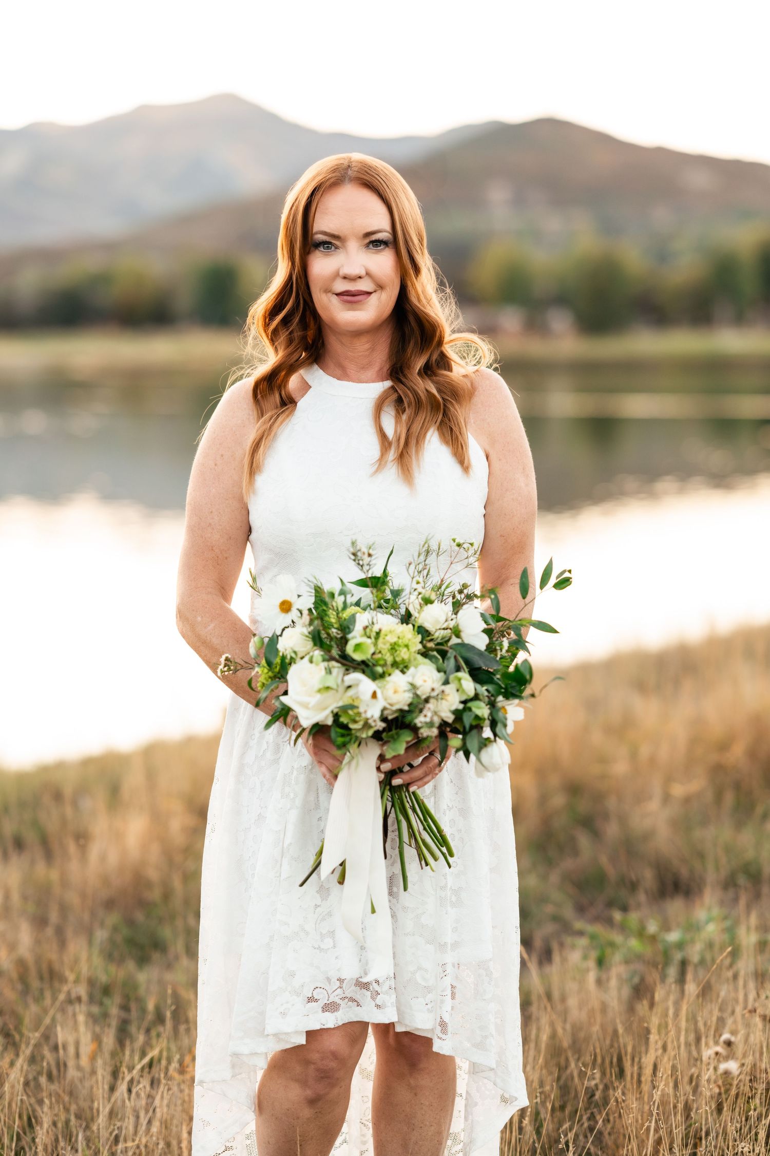 Bride poses with natural bouquet against mountain backdrop and golden field at sunset.