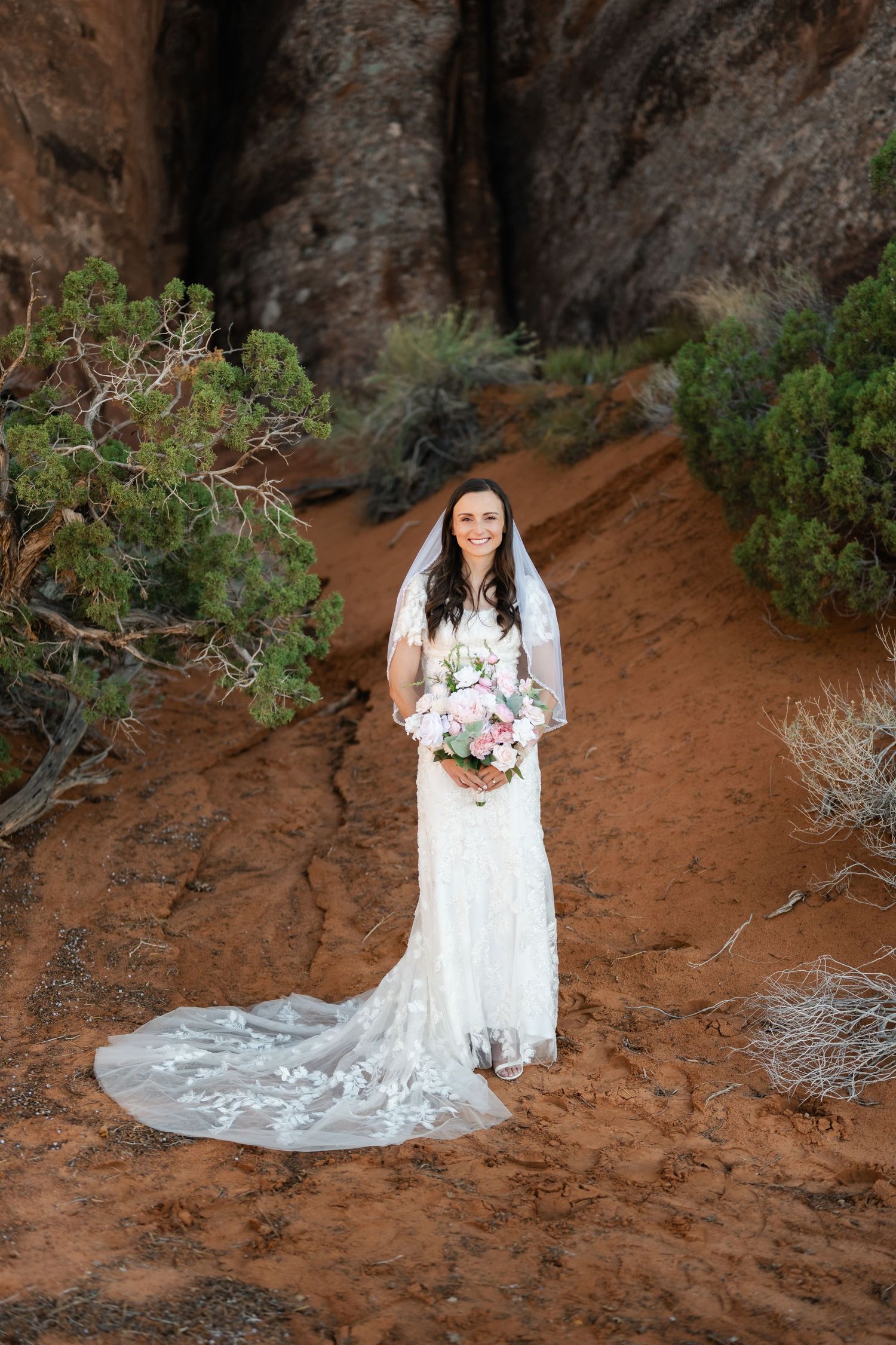 A bride in a lace wedding gown with long train stands among desert flora and red rock formations.
