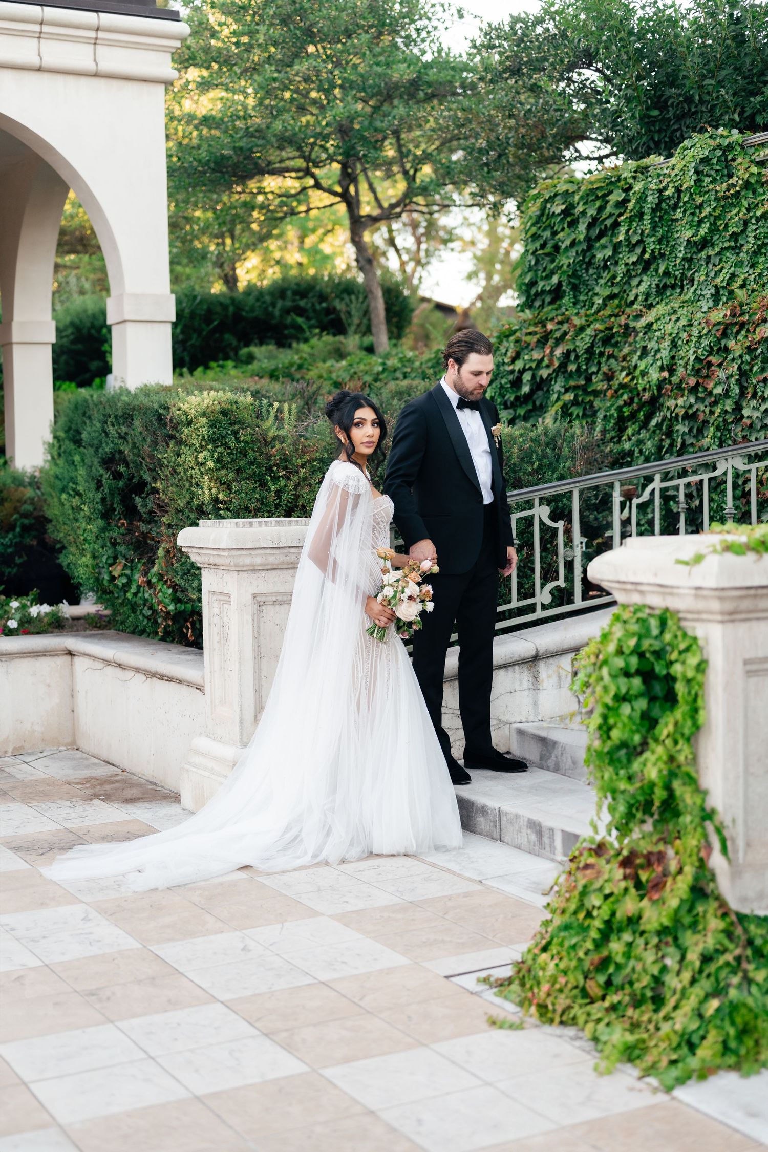 A bride and groom share a private moment on elegant white stone steps surrounded by lush greenery at an outdoor venue. UVU Museum of Art